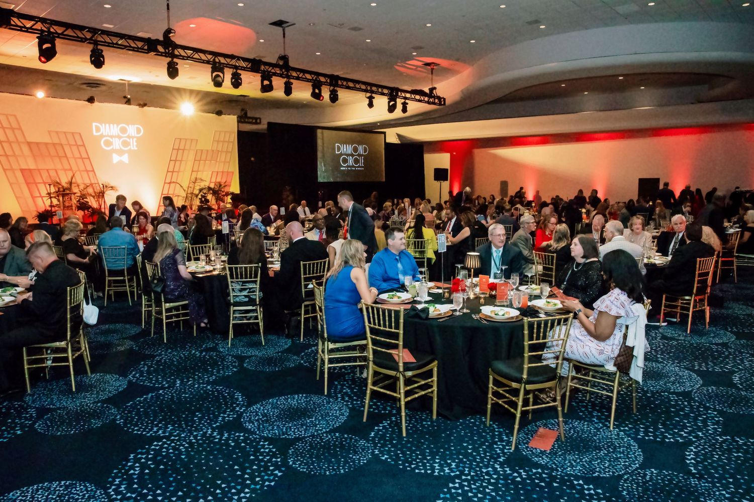 Guests dine at round tables in a large ballroom during a formal gala event with stage lighting.