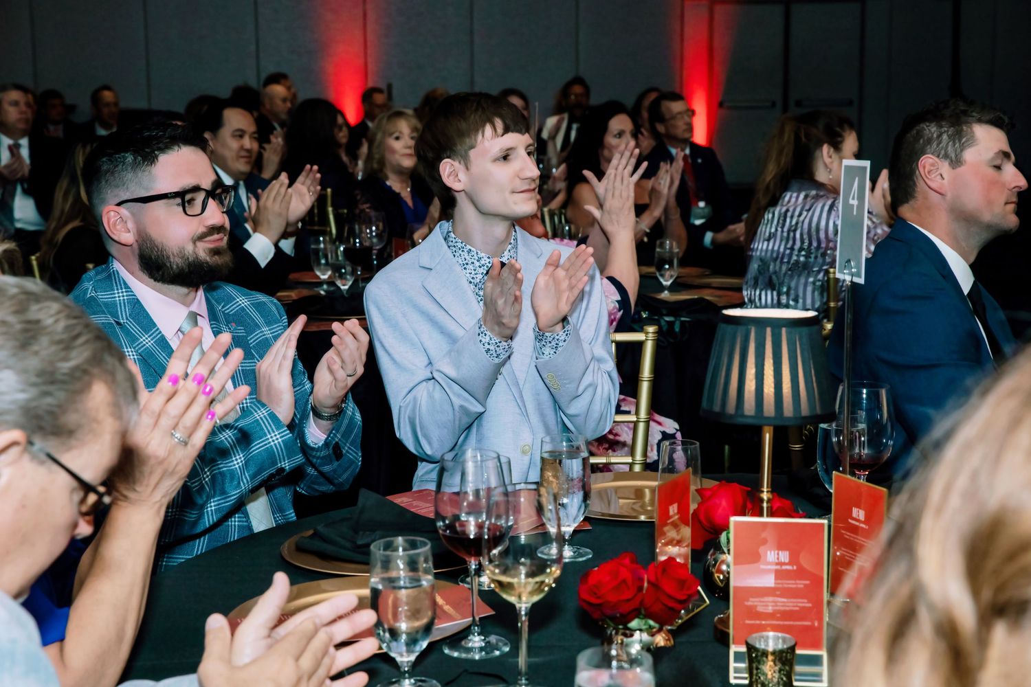 Guests applaud at an elegant gala dinner event with red roses, wine glasses, and ambient lighting.