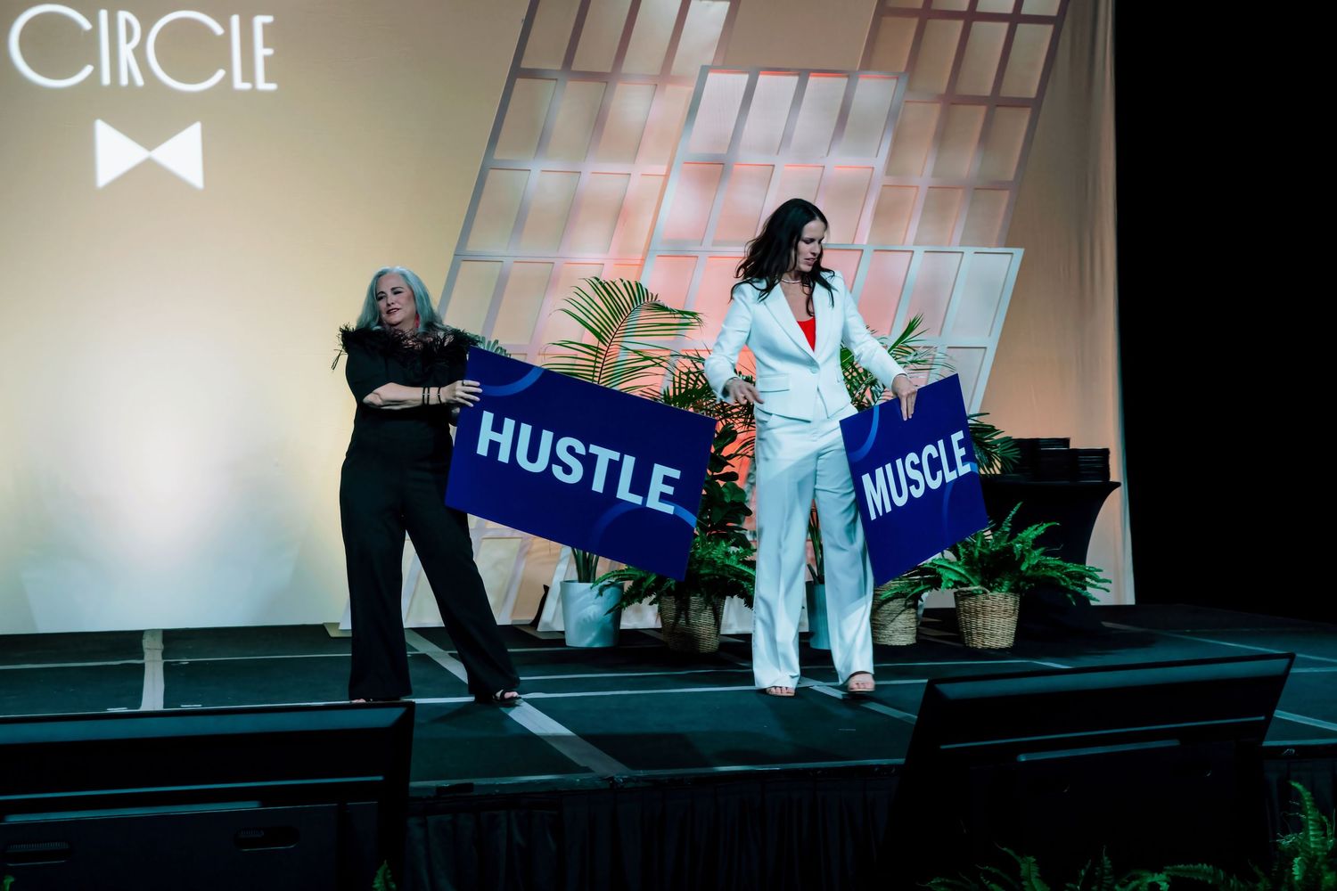 Two women on stage holding Hustle and Muscle signs at Circle event.