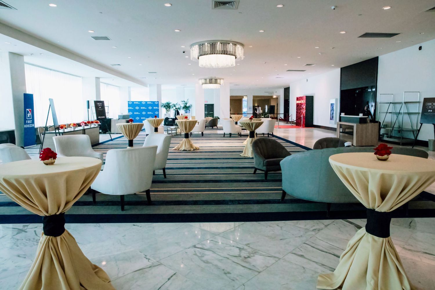 Elegant hotel lobby with draped cocktail tables, white lounge chairs, marble floors, and chandelier lighting for events.