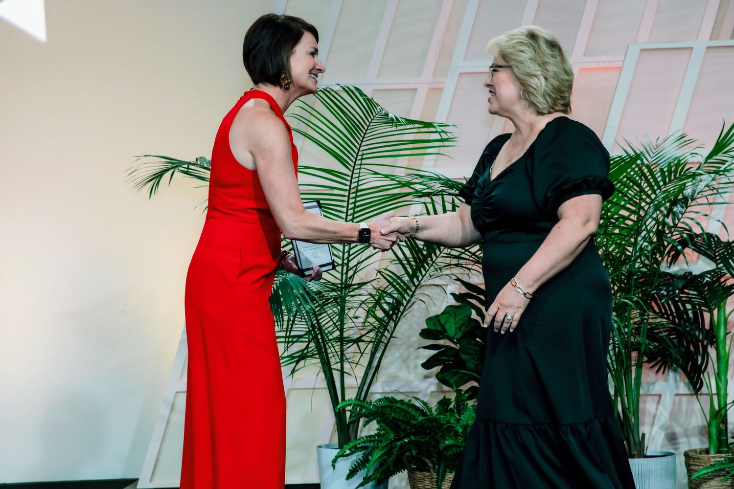 Two women in elegant dresses shaking hands at a formal event surrounded by tropical plants.