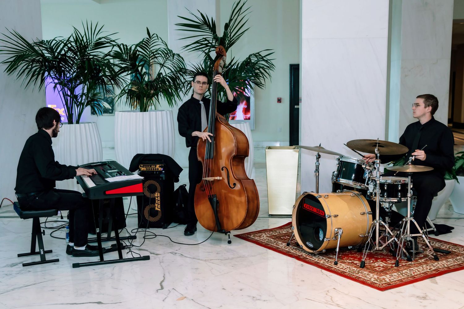 A jazz trio performs with keyboard, upright bass, and drums in an elegant white lobby with tropical plants.