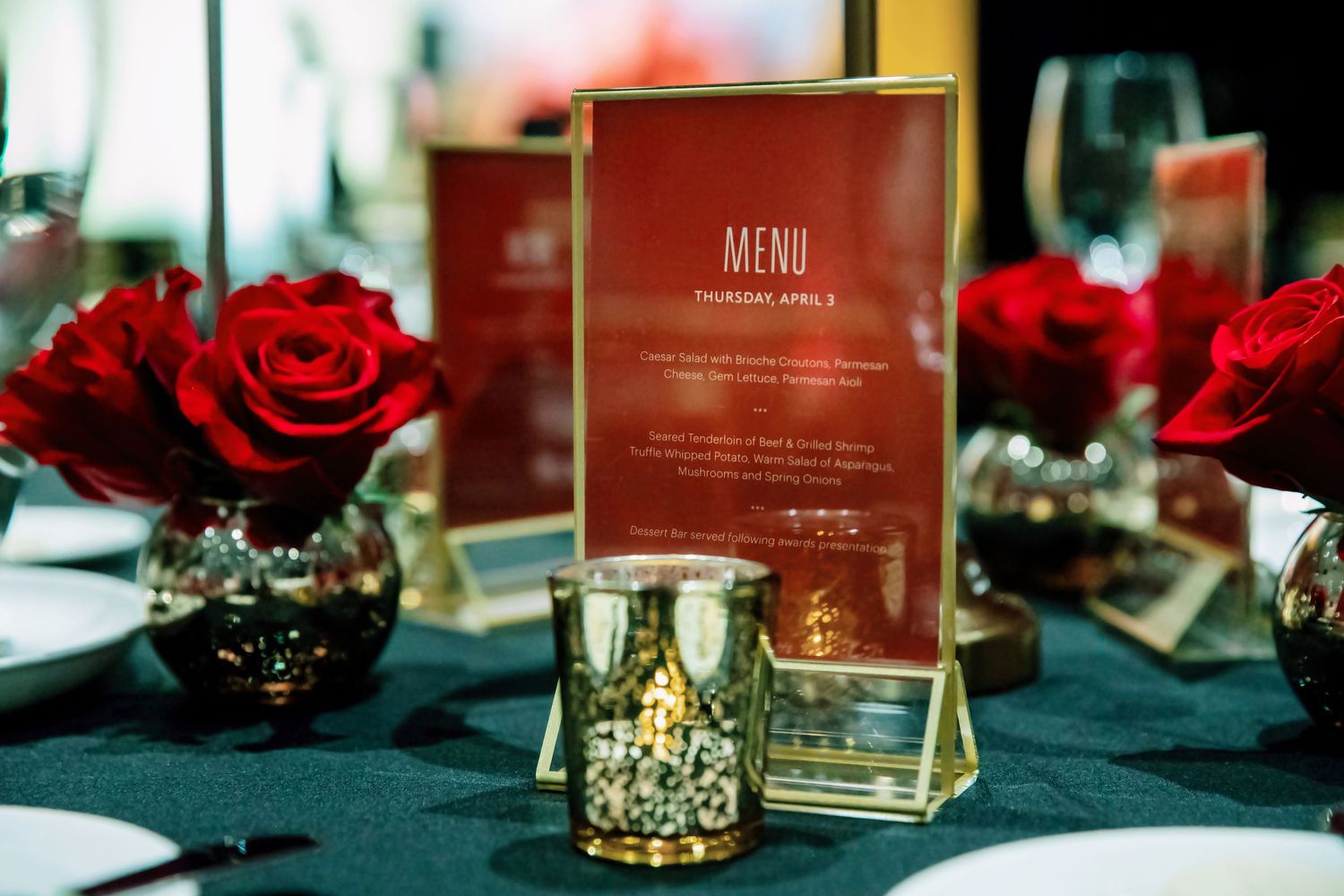 Elegant event table setting with red roses, gold candle holders, and a red menu card for a formal dinner.