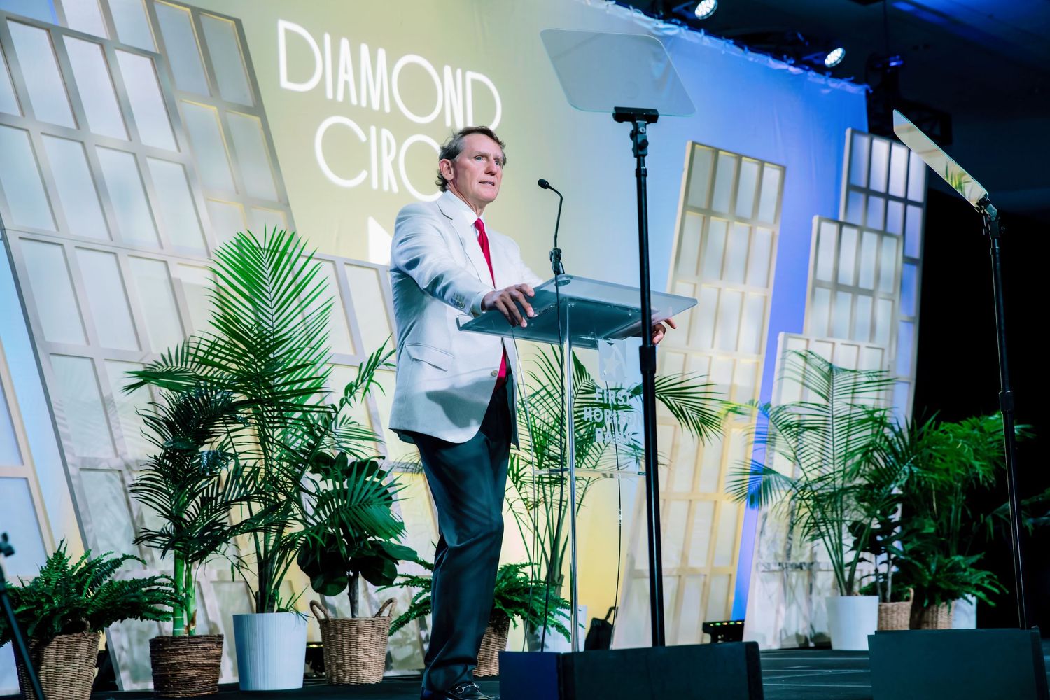 Man in white blazer speaks at Diamond Circle event podium, surrounded by tropical plants on an elegant stage.
