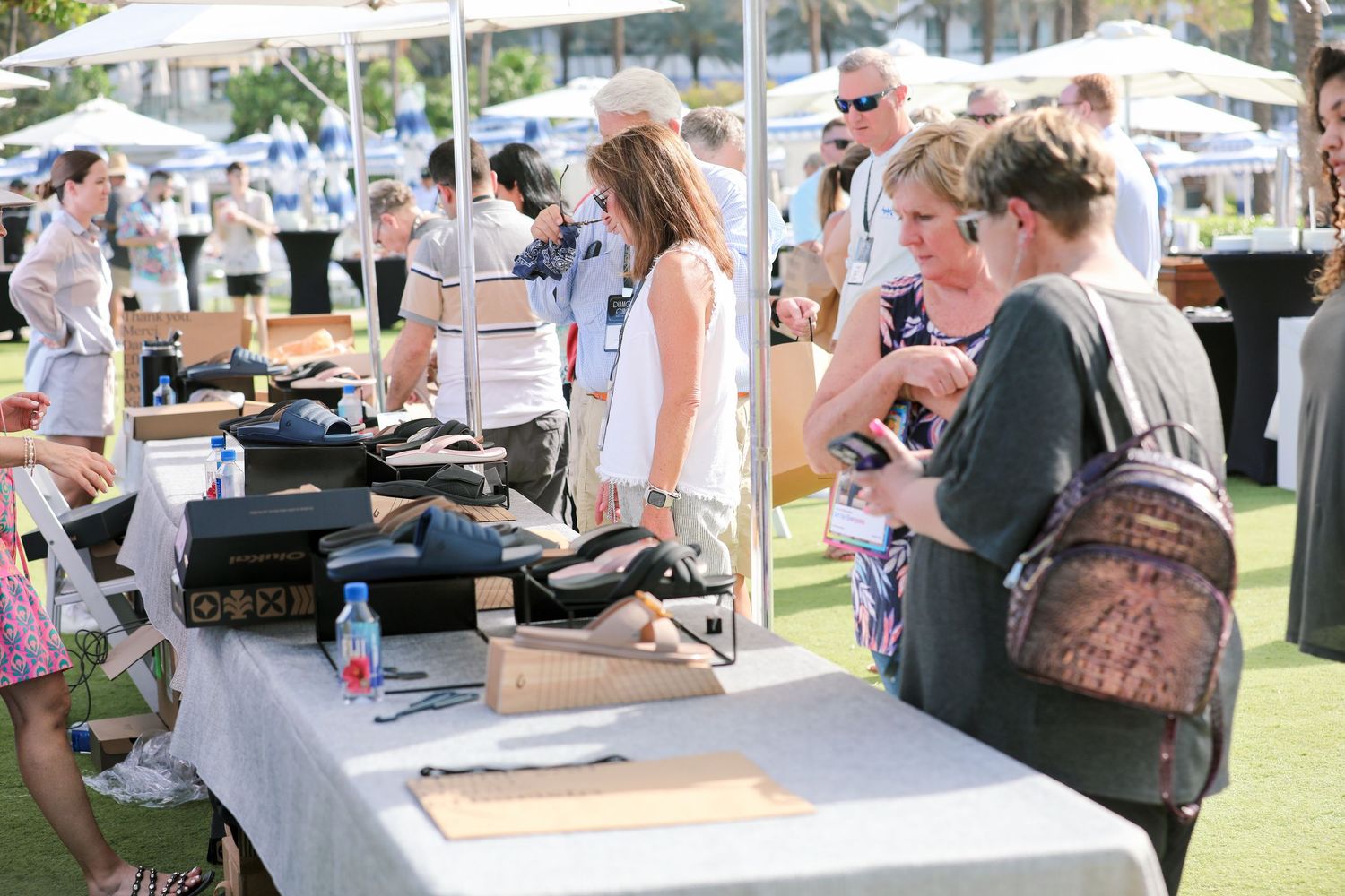 Shoppers browse handmade goods at an outdoor market vendor table on a sunny day.
