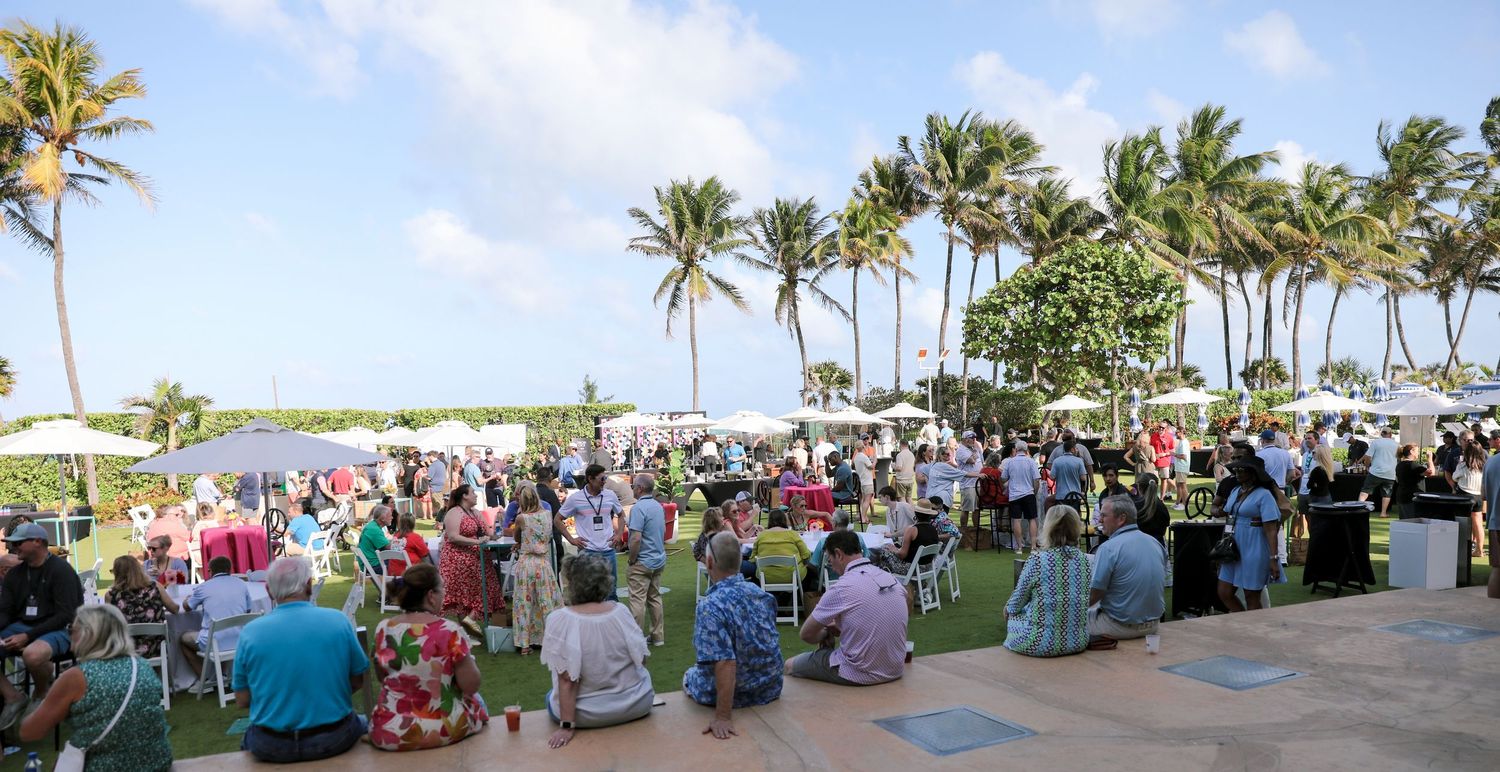 Outdoor gathering with crowd seated on lawn near palm trees at a tropical beachside event under blue skies.