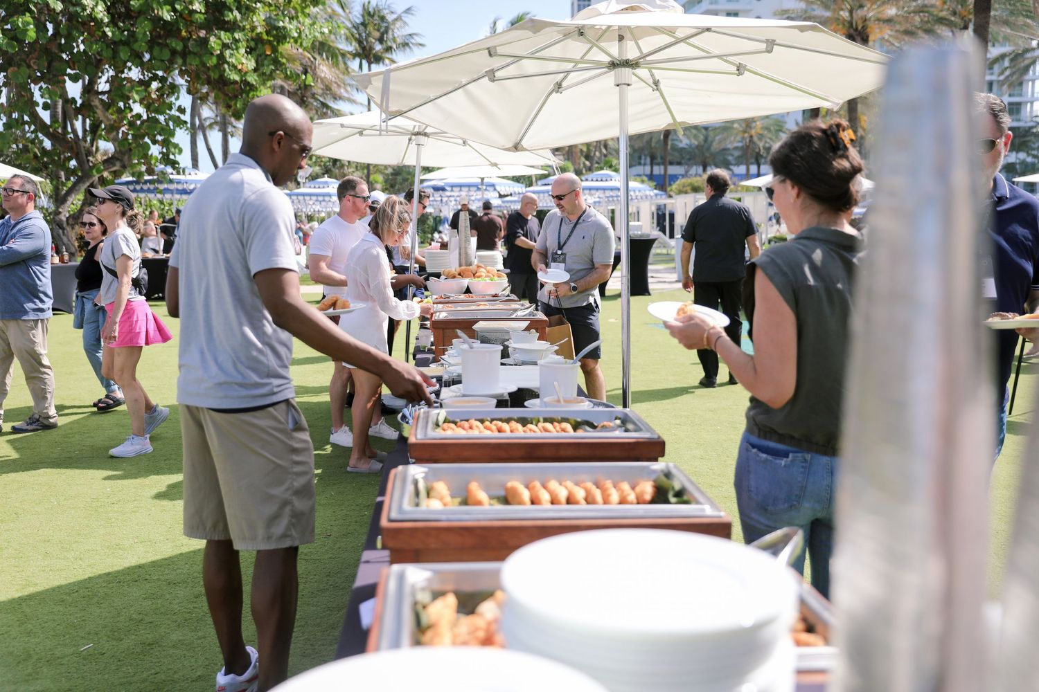 Guests enjoy an outdoor buffet spread under white umbrellas at a sunny Miami event.