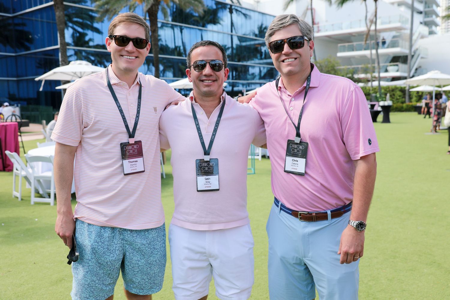Three men in pink polo shirts wearing lanyards smile together outdoors at a sunny golf event.