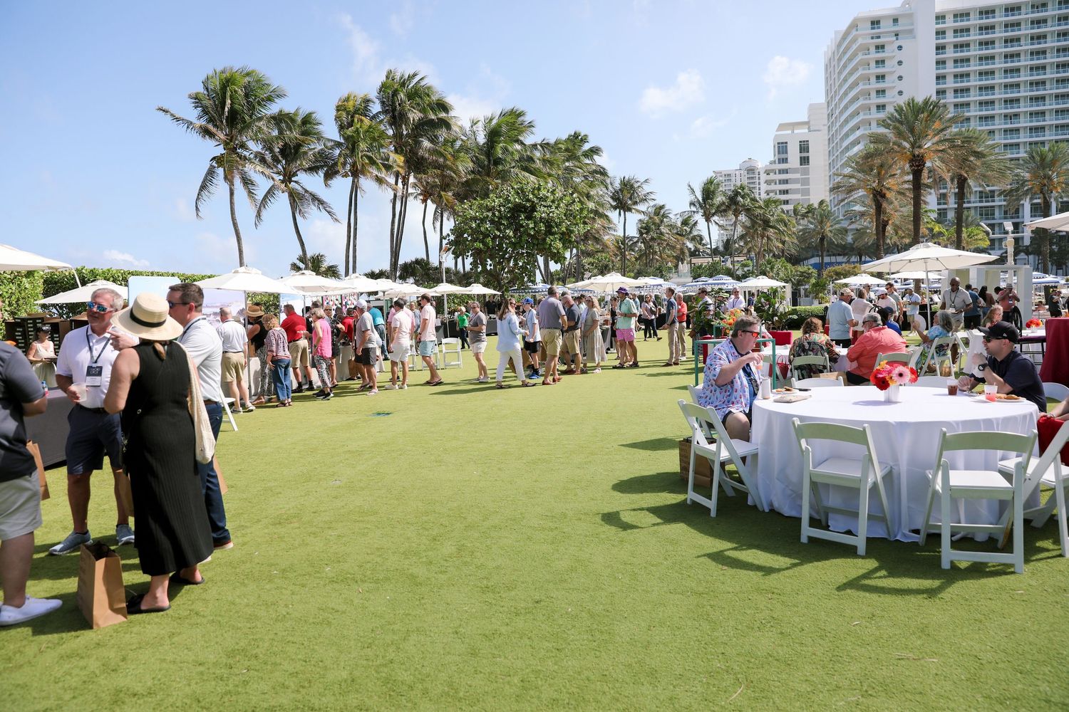 Outdoor event on lush green lawn with crowds, white tents, palm trees, and a luxury hotel in Miami Beach.