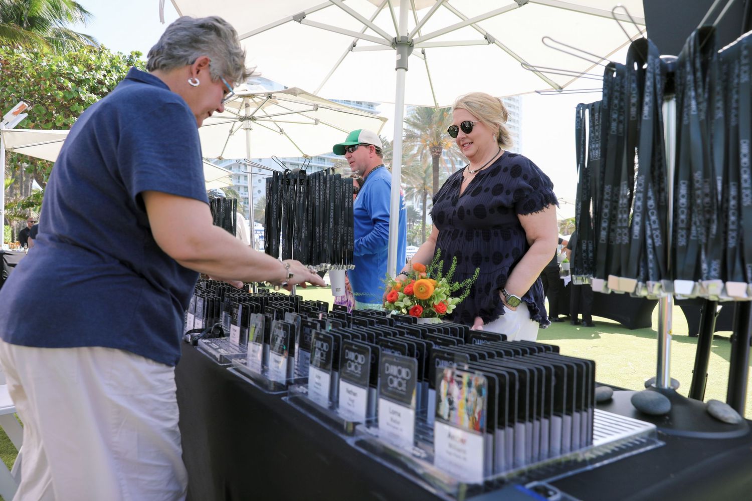 Outdoor vendor booth displaying rows of black merchandise under a white tent with two women browsing products.