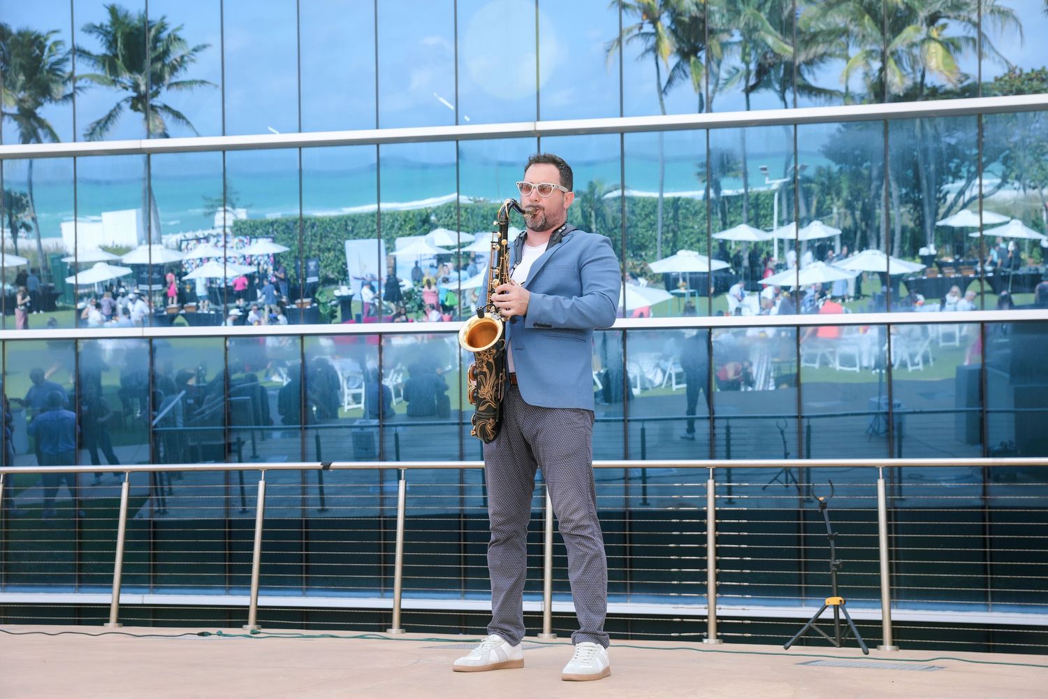 Saxophonist performs in grey suit outside glass building at Miami outdoor event.