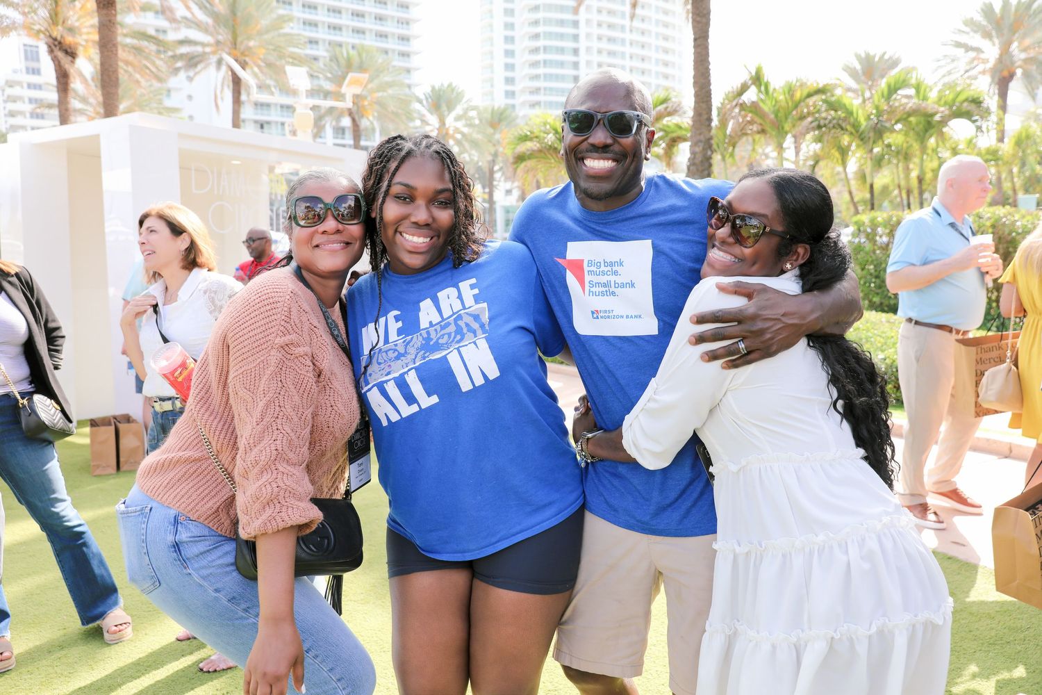 Four friends smile and hug outdoors at a Miami charity event on a sunny day.