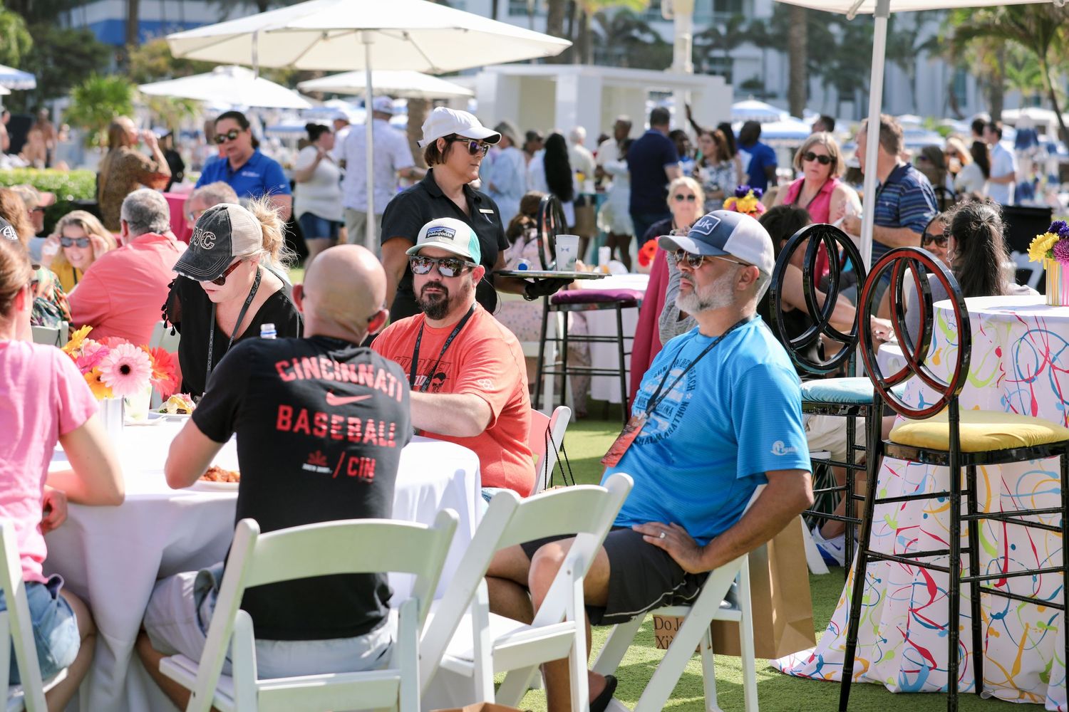 People sitting at white folding chairs and tables outdoors at a sunny community event or festival gathering.