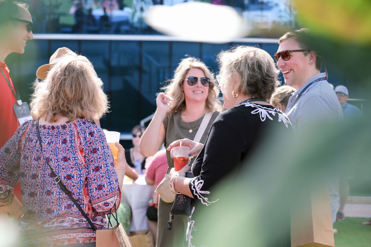 Group of people socializing outdoors at a sunny event, smiling and holding drinks in a festive gathering.