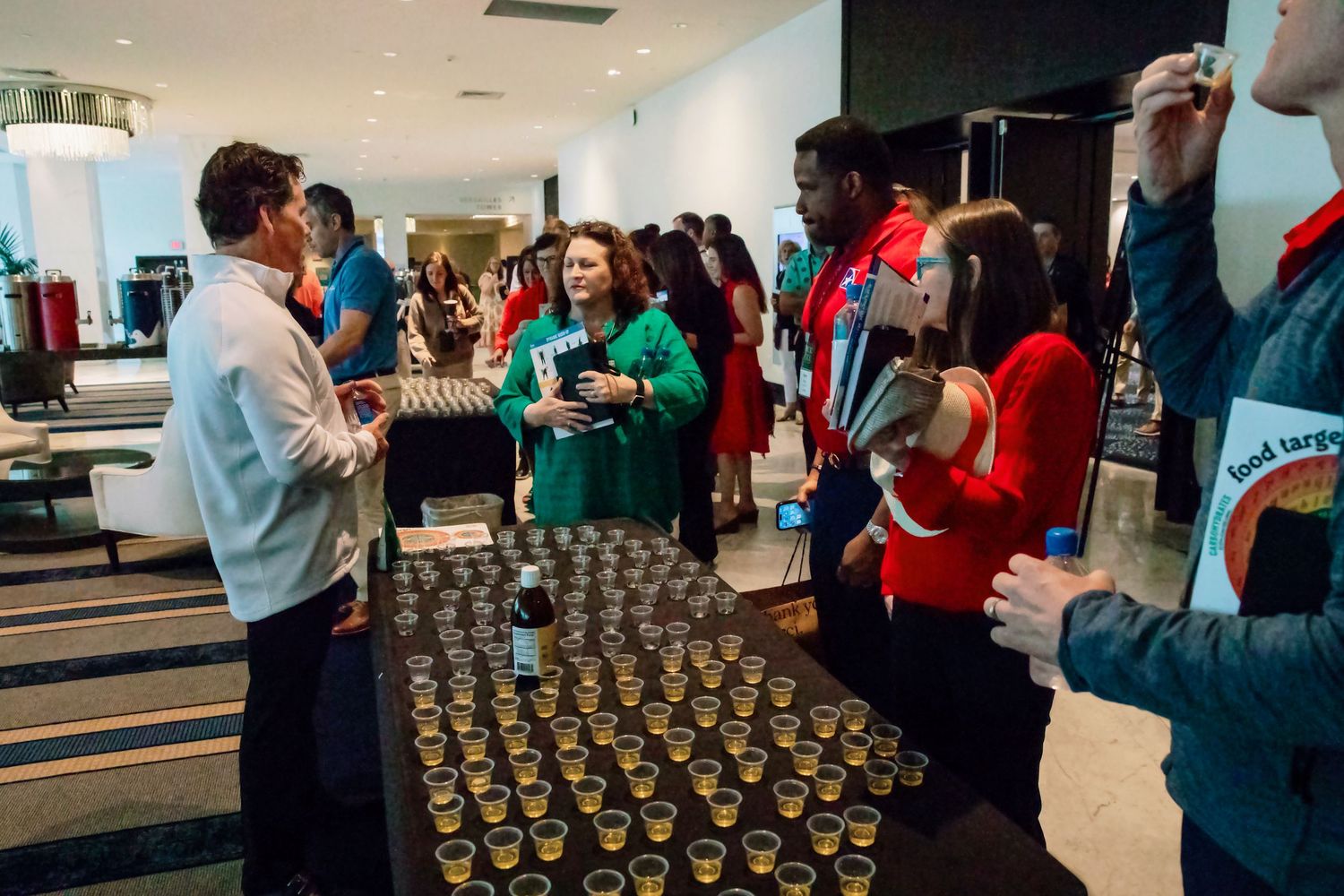 Attendees network and sample drinks at a corporate event reception with rows of small cups on display.