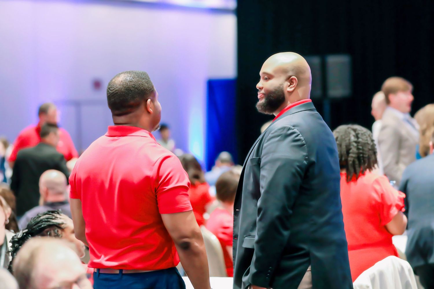 Two men in red and black attire converse at a formal event in a crowded banquet hall.