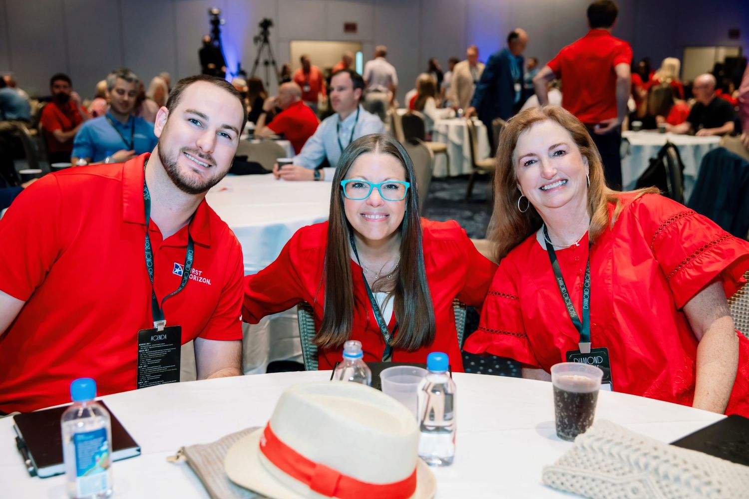 Three smiling professionals in red shirts pose together at a conference table during a corporate event.