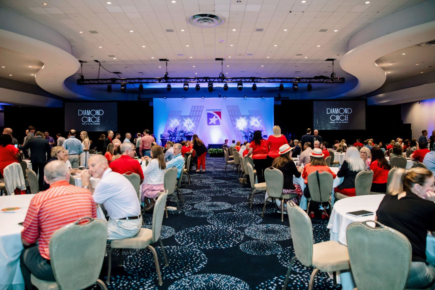Large conference banquet hall with guests seated at round tables facing a lit stage with blue backdrop.