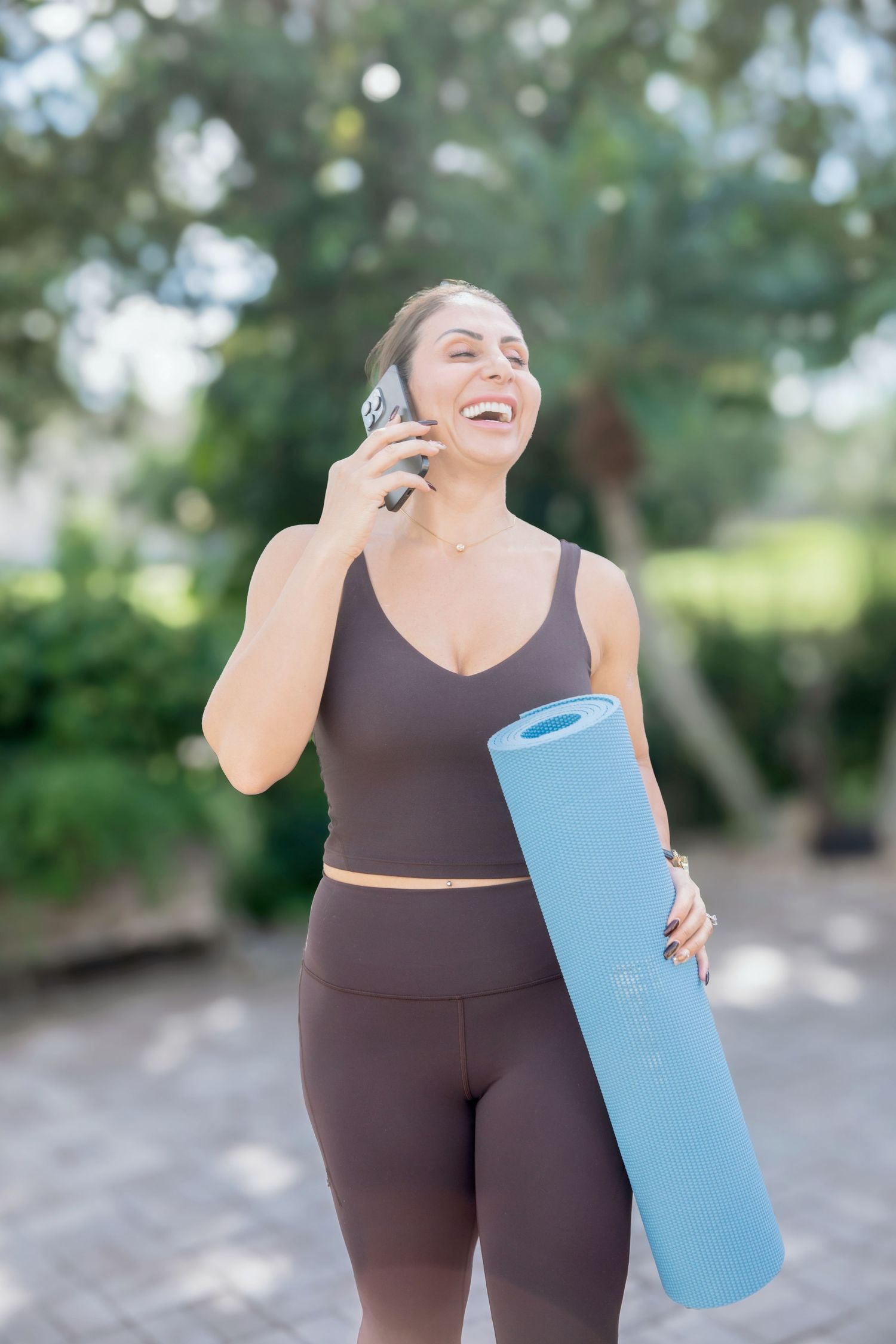 Smiling woman in brown activewear holds a blue yoga mat while talking on her phone outdoors.