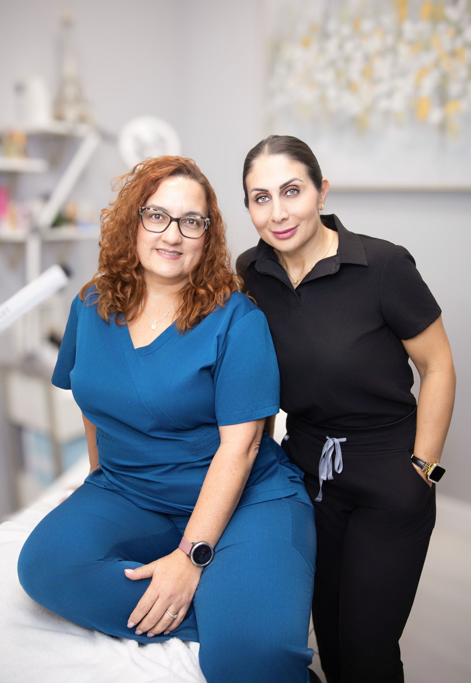 Two women in a medical spa setting, one in blue scrubs and one in black, smiling warmly at the camera.