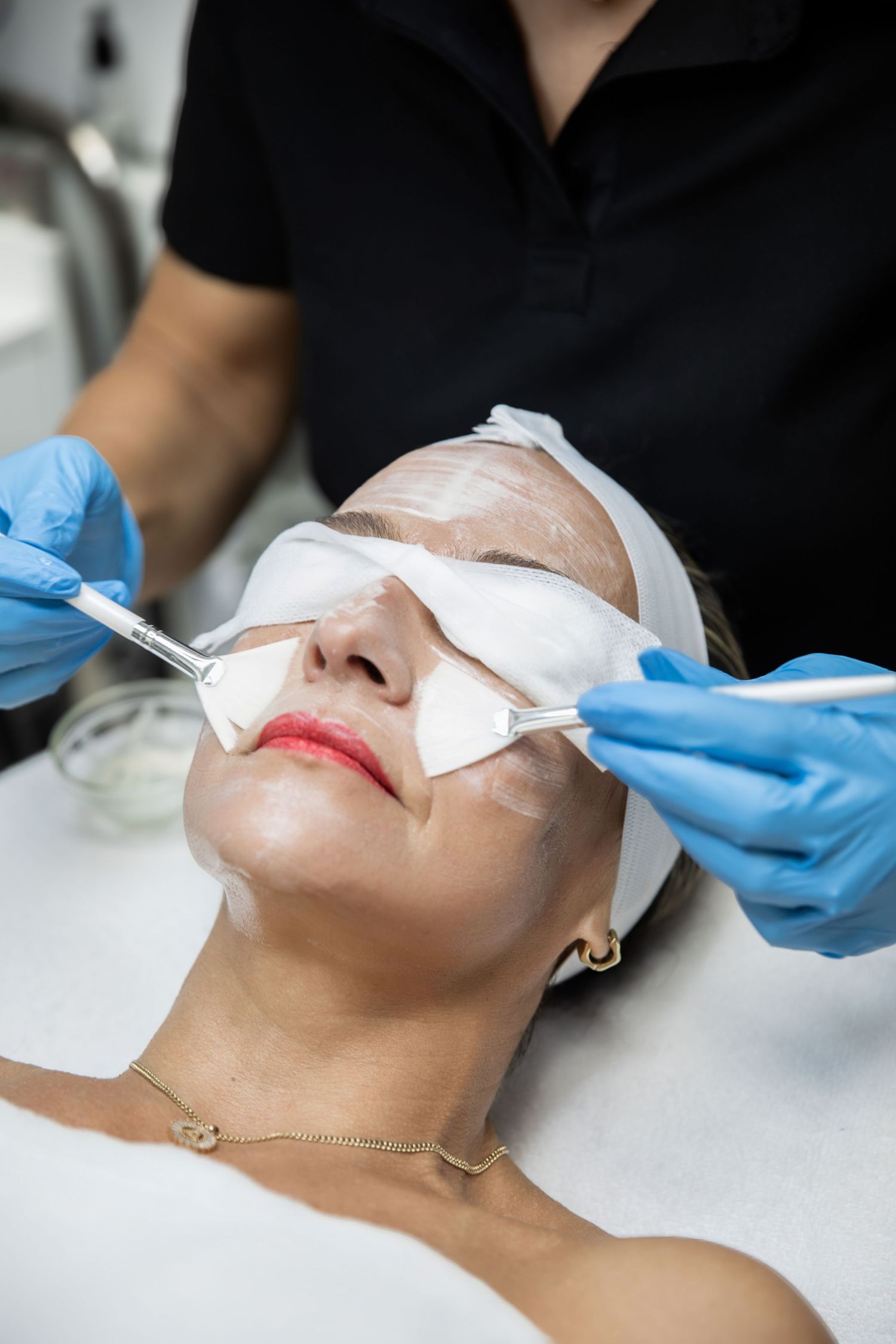 Esthetician applying white facial mask with brushes on woman lying on treatment table in beauty salon.