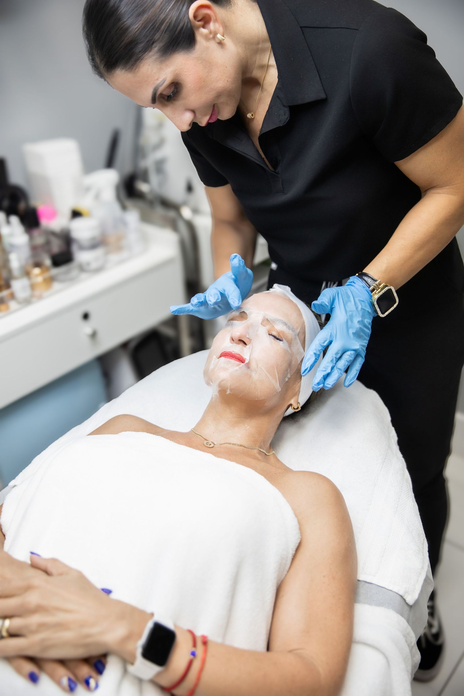 Esthetician in black uniform applying facial sheet mask to mature woman lying on treatment table in spa.