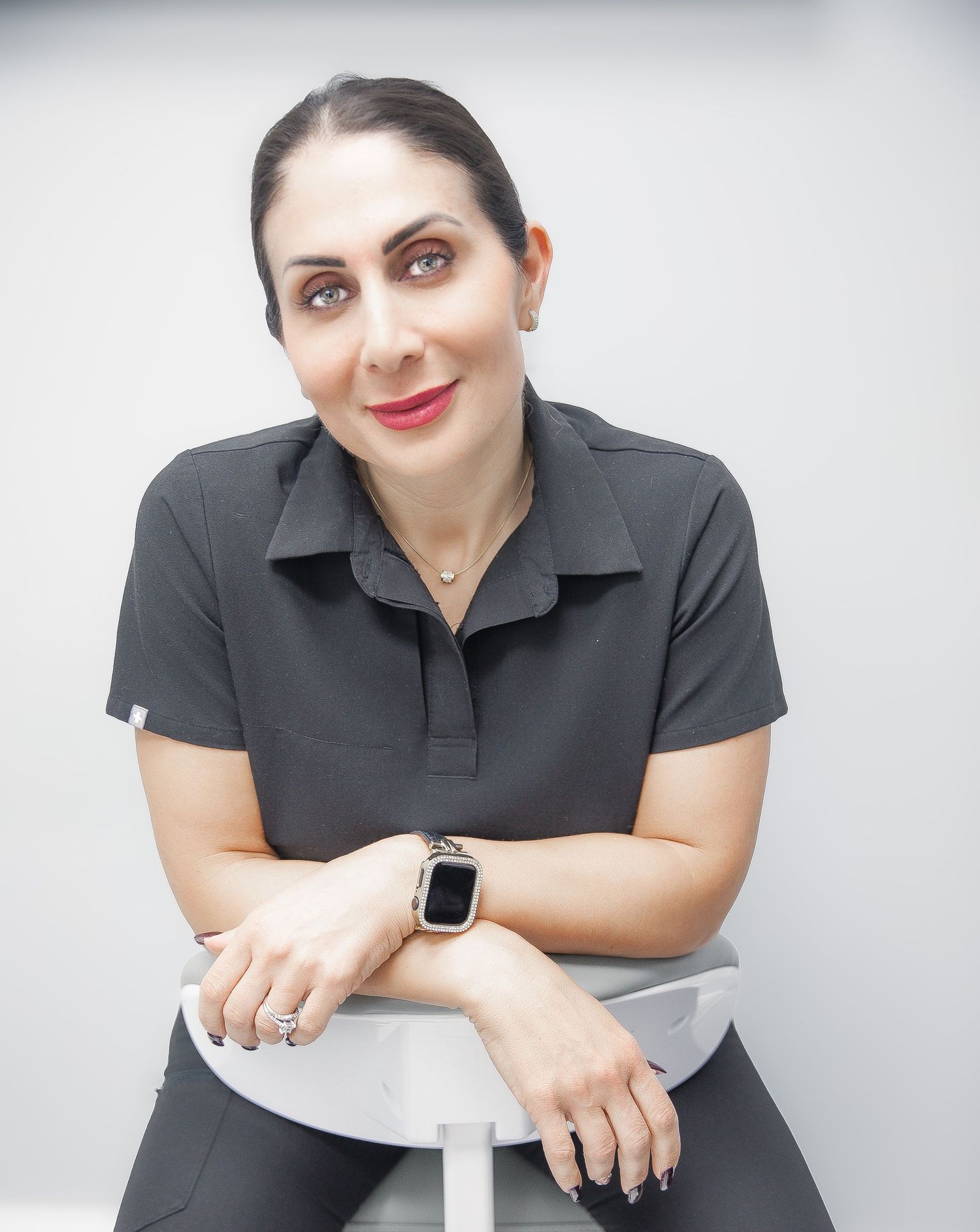 Woman in dark polo shirt sitting relaxed on stool, wearing smartwatch and rings, smiling confidently.