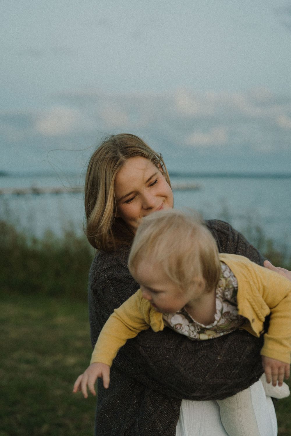 A smiling young woman holds a baby in a yellow outfit near a serene waterfront on a cloudy day.