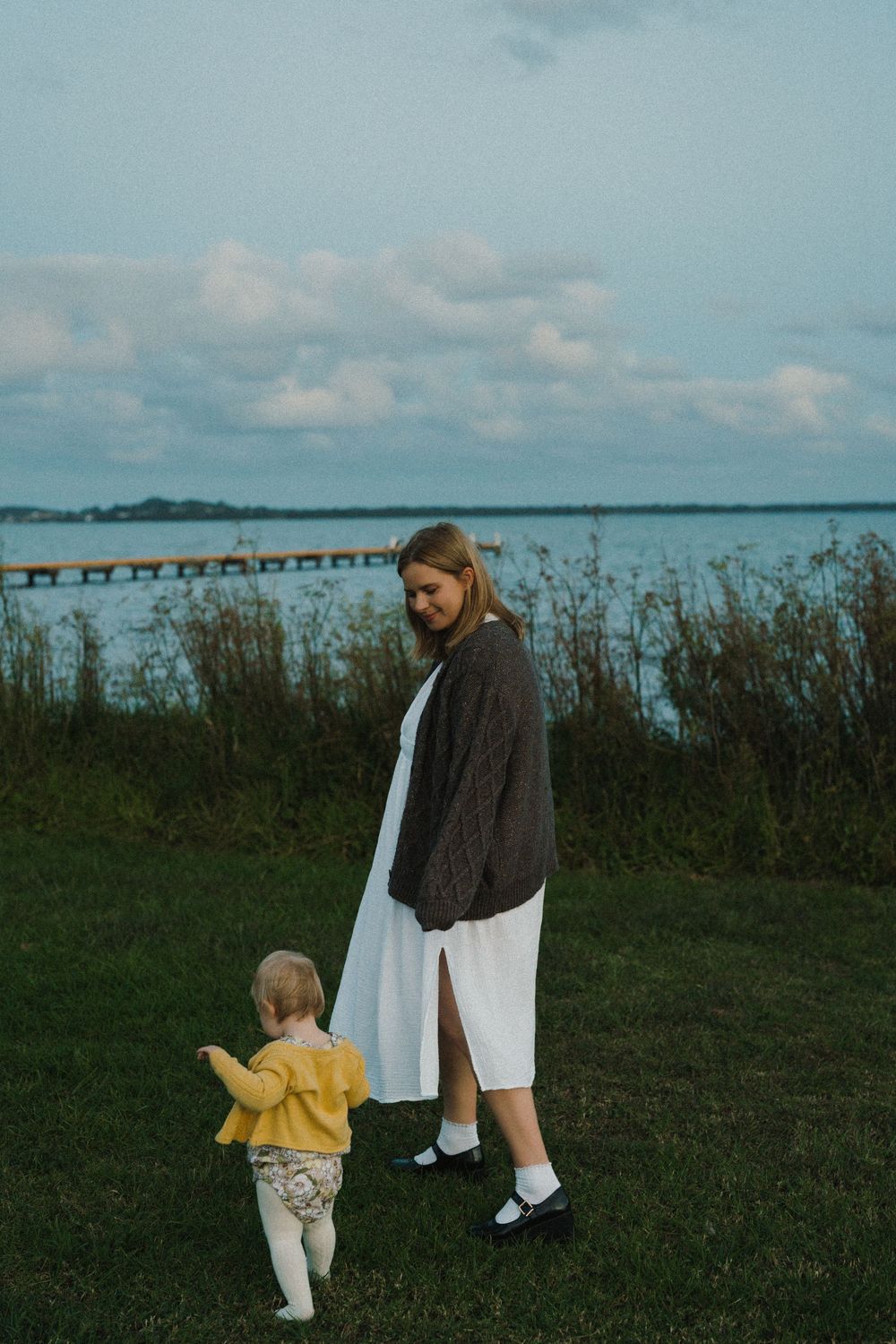 A mother in a white dress and gray cardigan walks with her toddler in a yellow sweater by a serene lakeside at dusk.