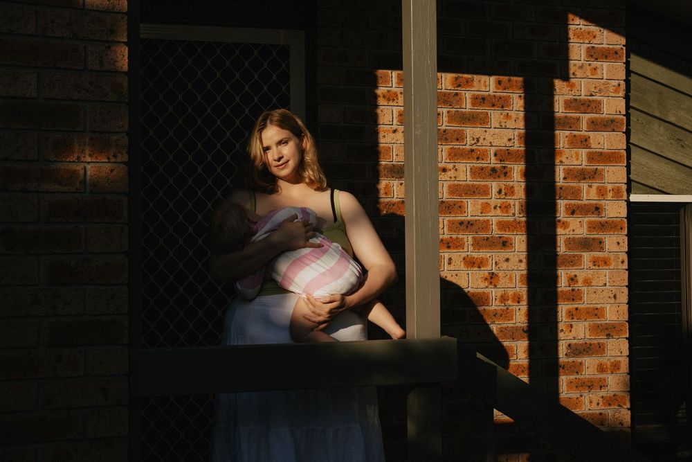 Pregnant woman sitting on a sunlit porch, cradling her baby bump, with warm light against a brick wall backdrop.