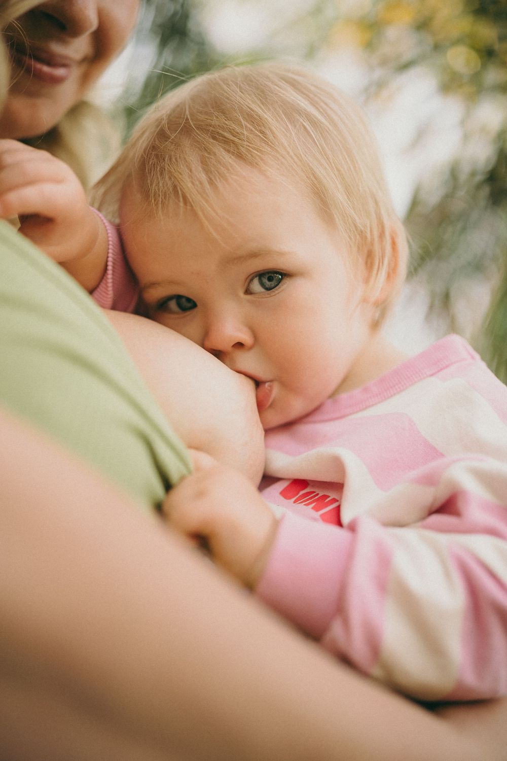 A blonde toddler in pink striped outfit being held and breastfed by a smiling mother outdoors.