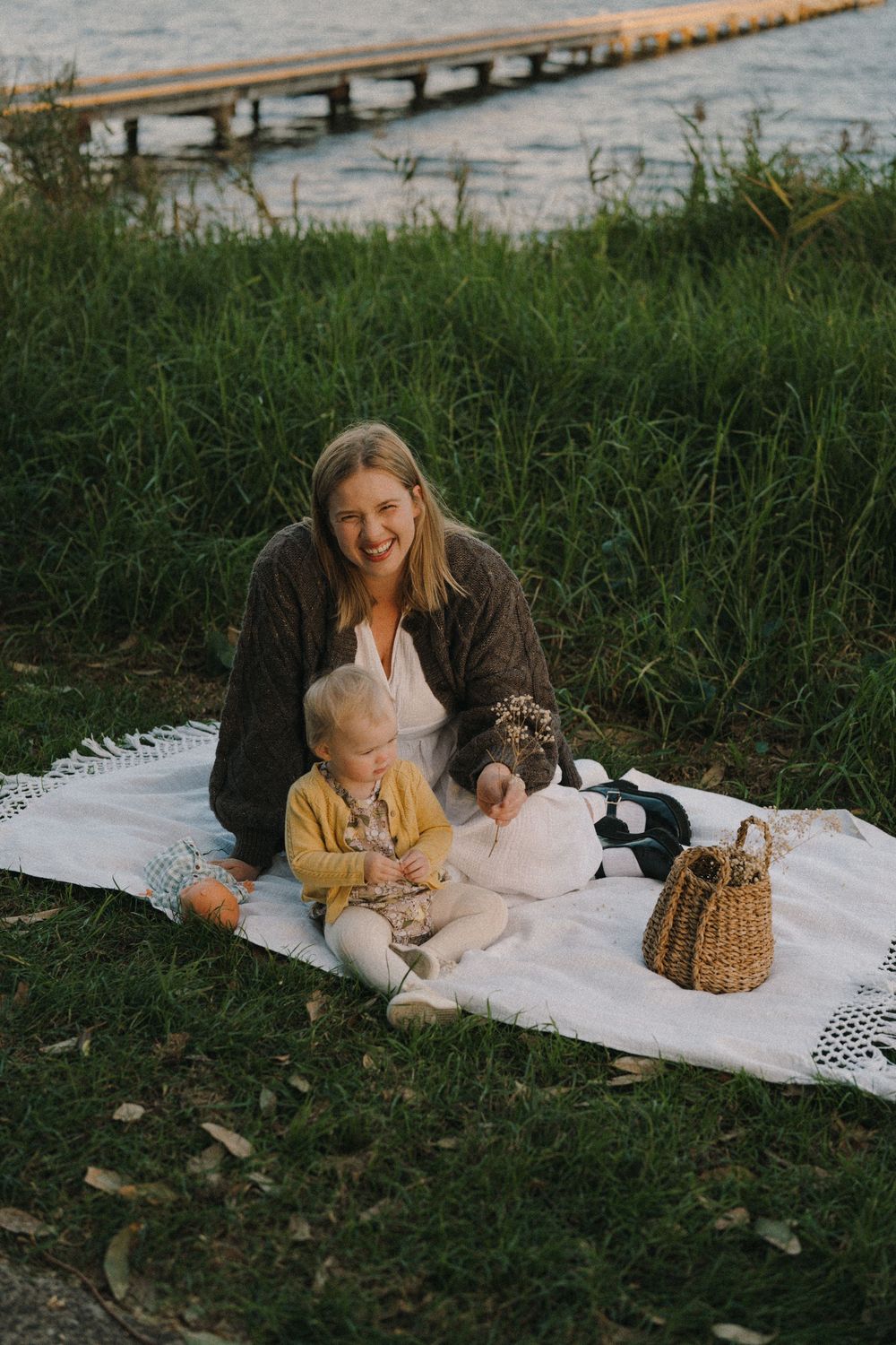 A smiling mother and toddler enjoy a lakeside picnic on a white blanket surrounded by lush green grass.
