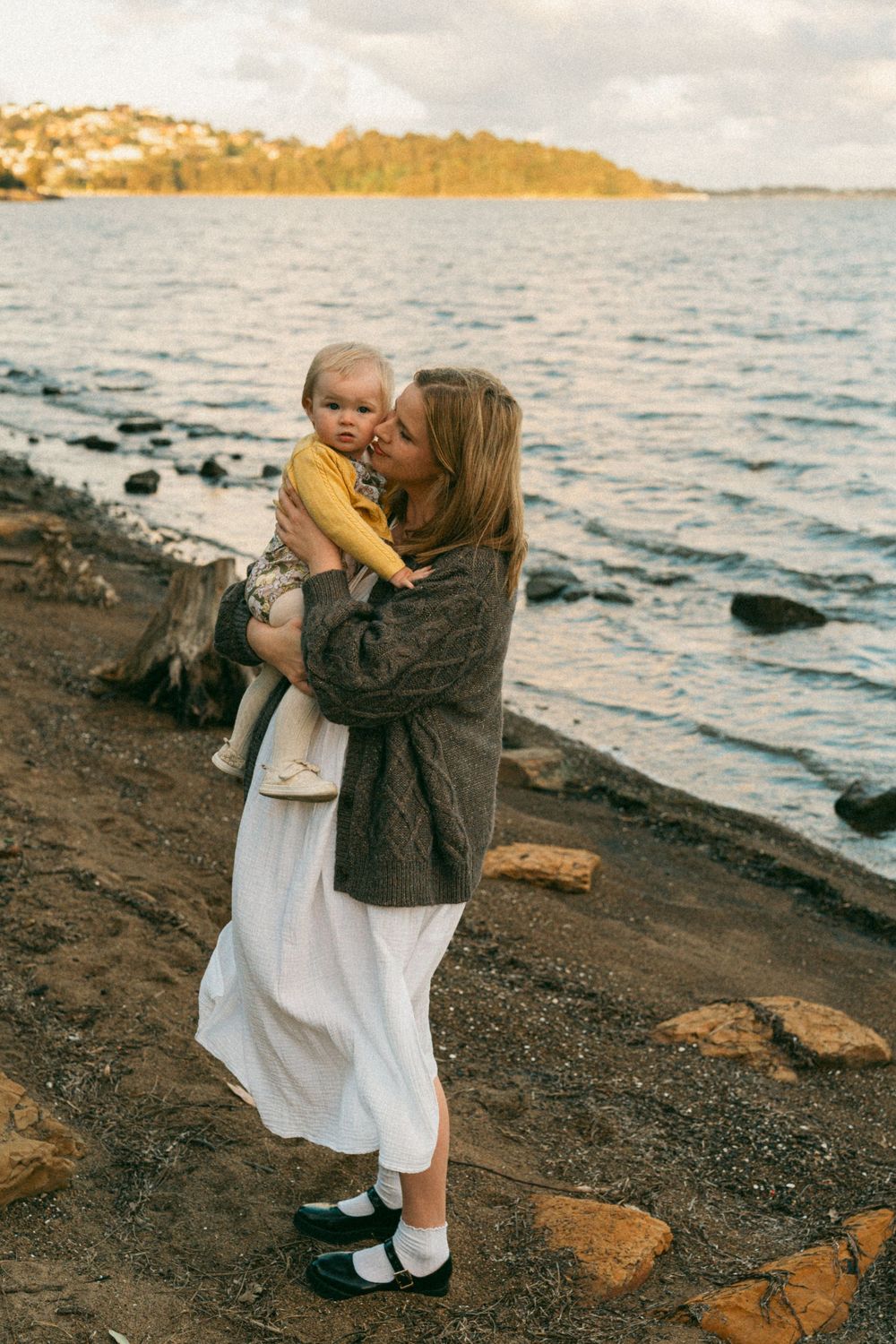 Woman in white skirt kisses baby by rocky shoreline at sunset.