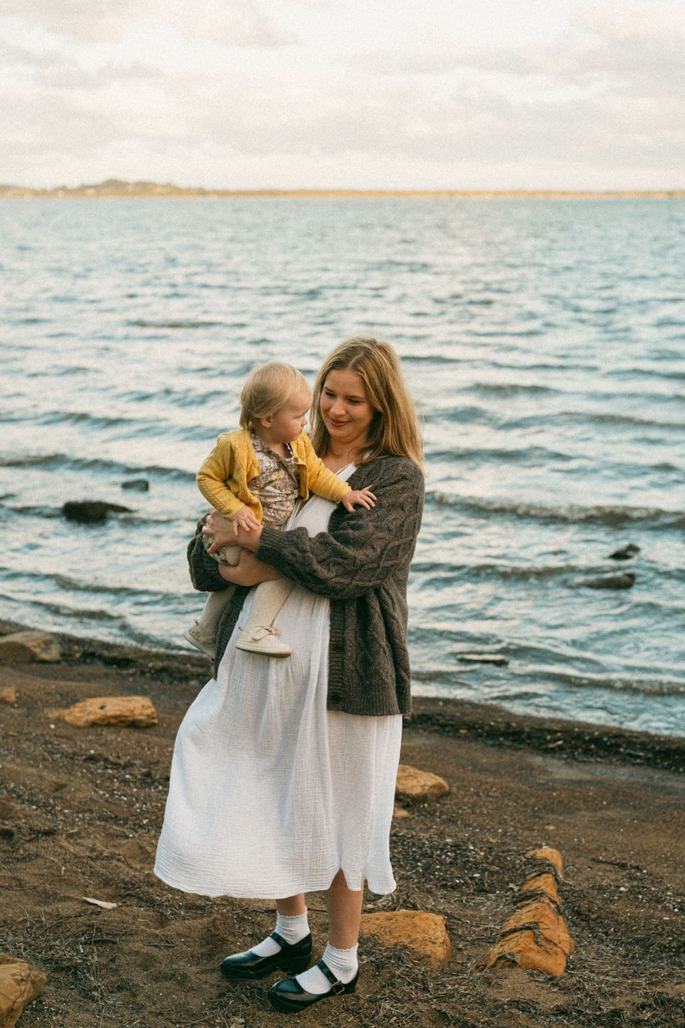 Mother holding young child on rocky shoreline with calm blue water in background on overcast day.