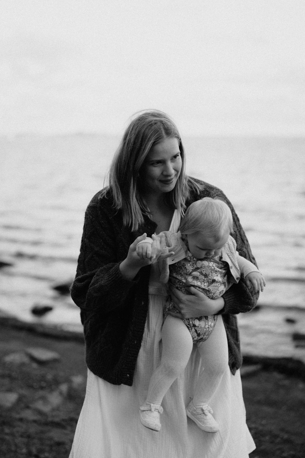 Black and white photo of a young woman holding a baby near a rocky shoreline, smiling warmly outdoors.
