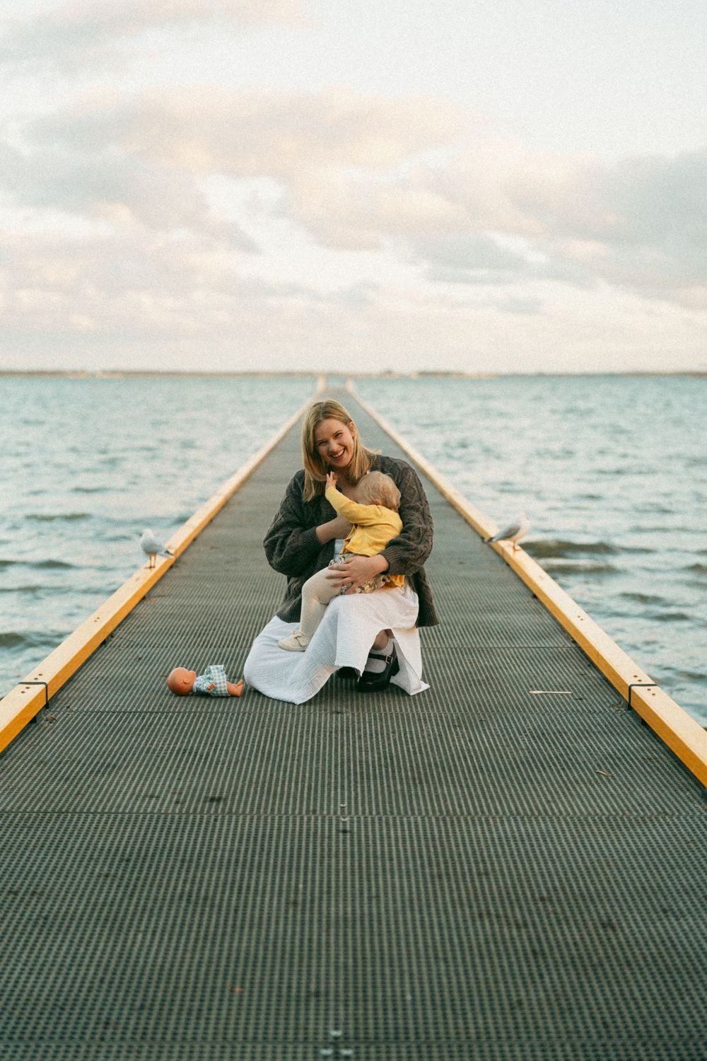 A smiling woman crouches on a pier holding a baby in a yellow outfit, with calm ocean waters and cloudy sky behind.