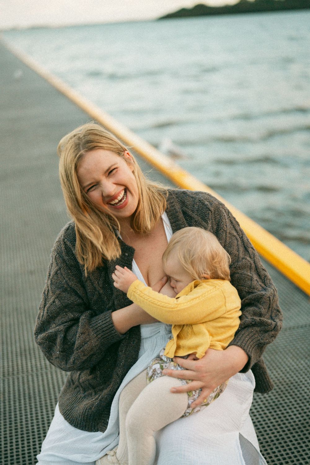 Smiling blonde mother breastfeeding toddler in yellow outfit on waterfront dock wearing brown knit cardigan.
