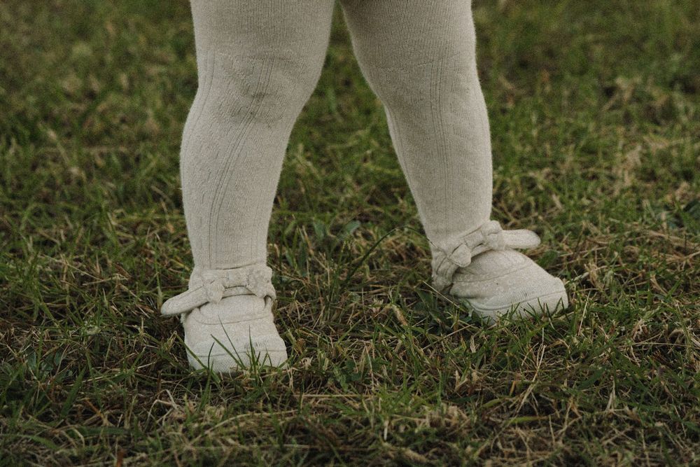 Close-up of toddler legs in white tights and white Mary Jane shoes standing on green grass.