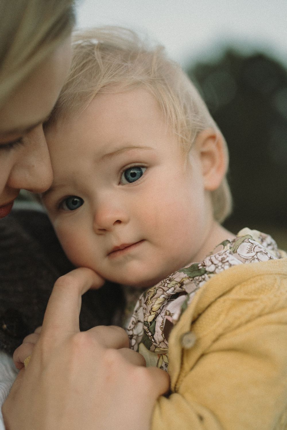 Close-up of a blue-eyed blonde toddler being held by a parent, wearing a yellow cardigan and floral bib.