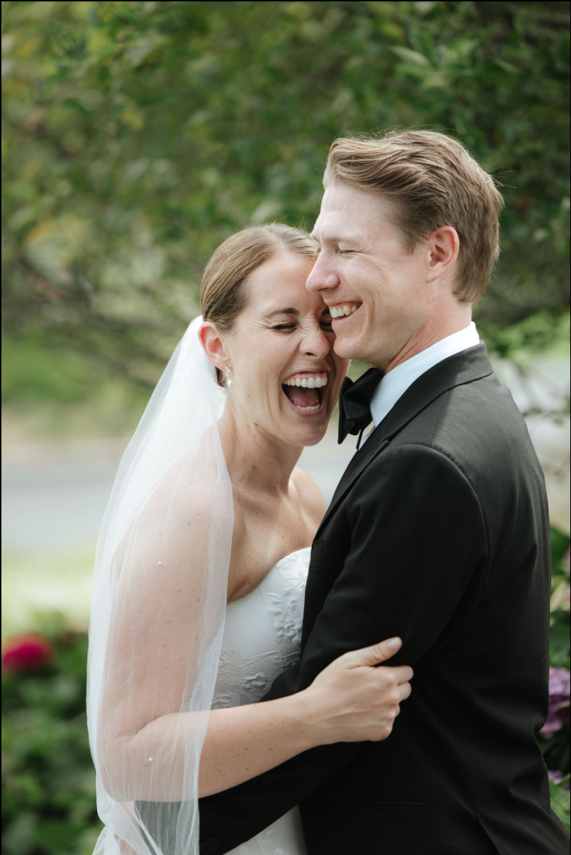 Joyful bride and groom laughing together outdoors, bride wearing veil and strapless gown, groom in black tuxedo.