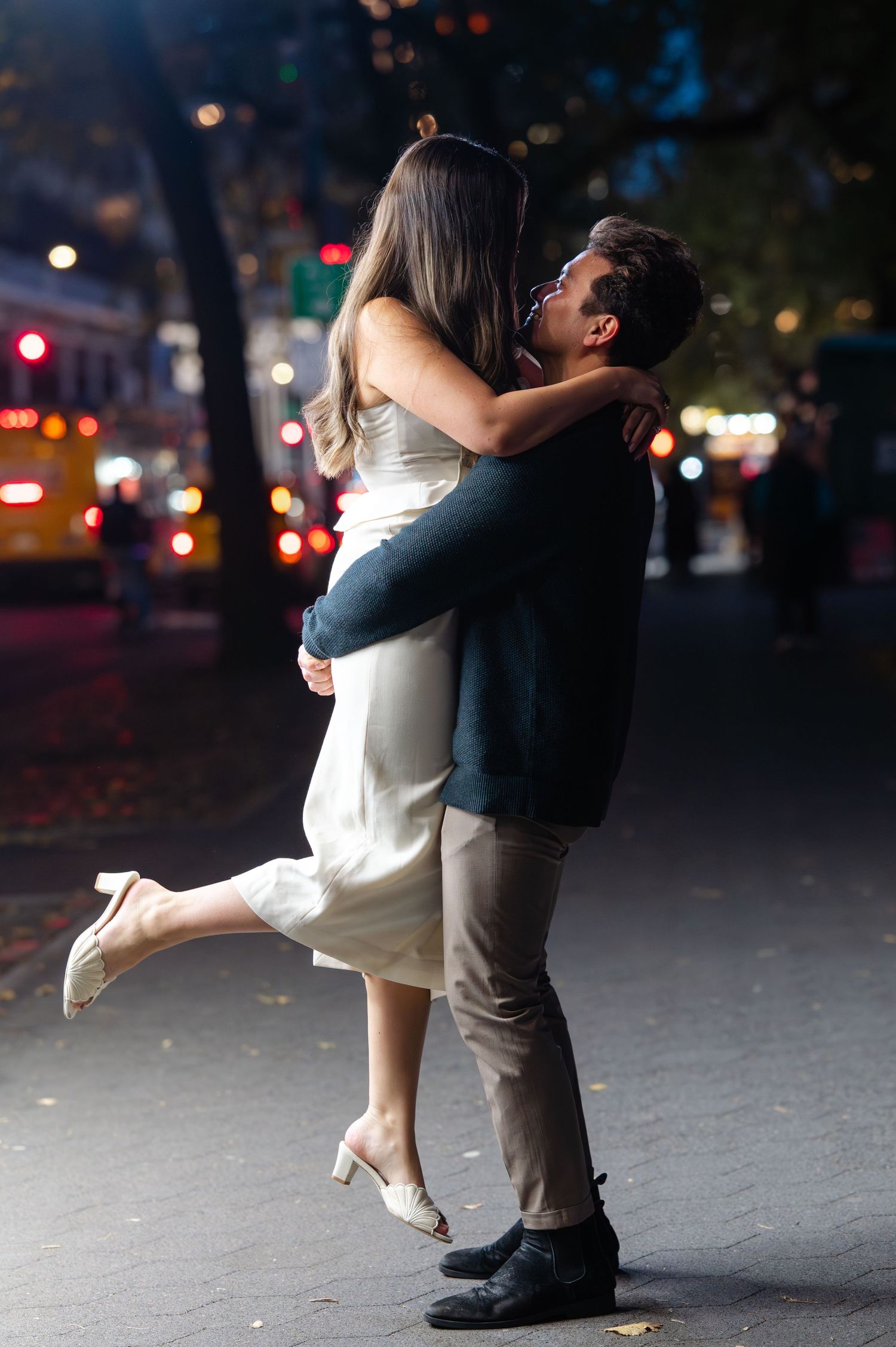 Couple embracing romantically on a city street at night, woman lifted off ground in white dress and heels with bokeh in the back.