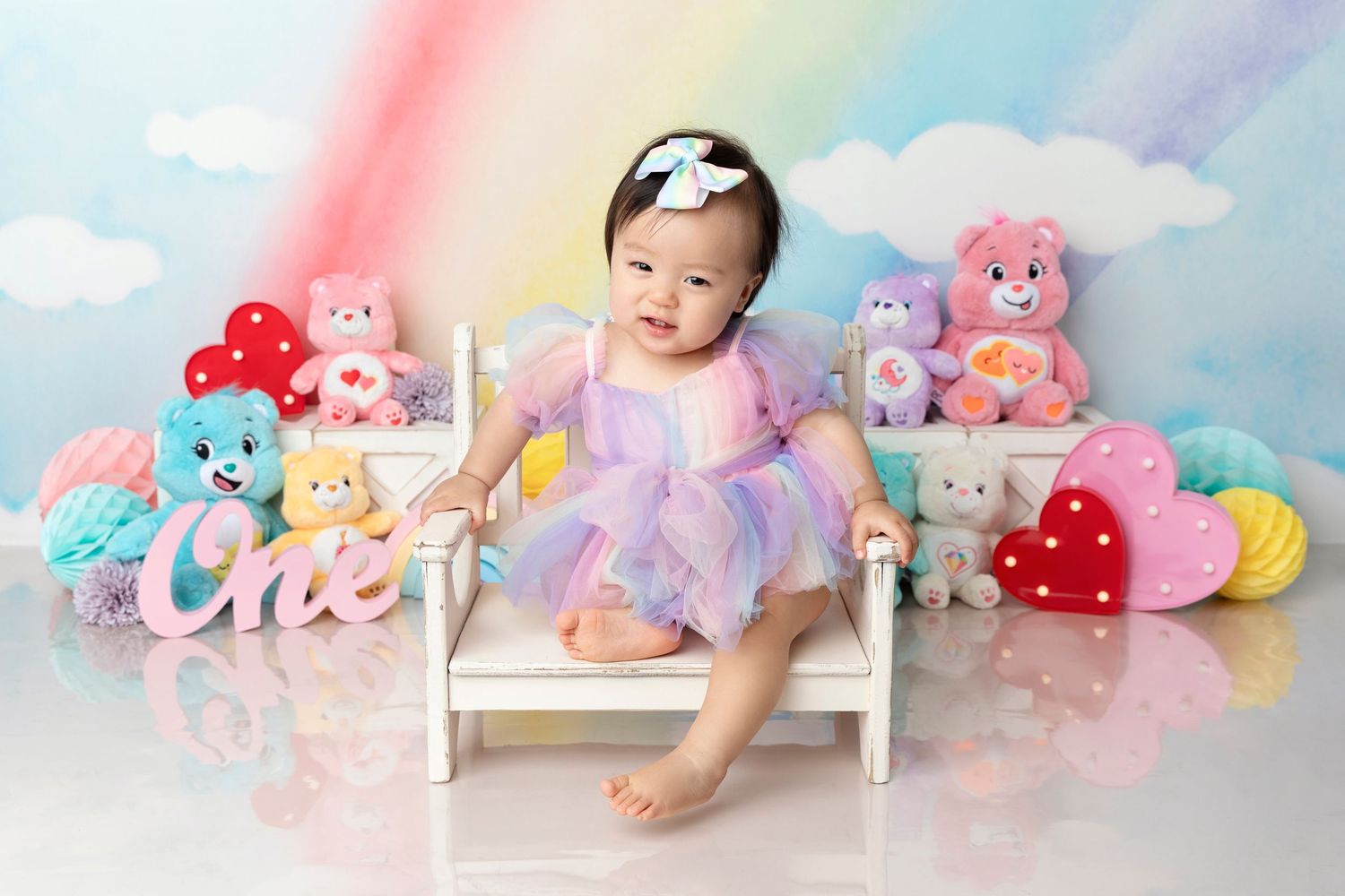 Baby girl in purple tutu sits among Care Bears and hearts with a rainbow backdrop celebrating her first birthday.