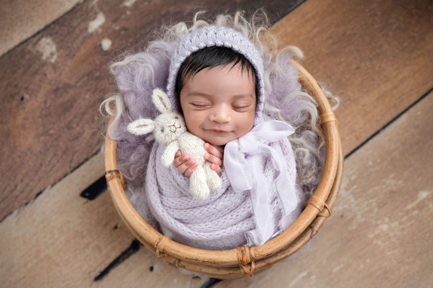Sleeping newborn baby wrapped in lavender swaddle with bunny toy in wicker basket on wood floor.