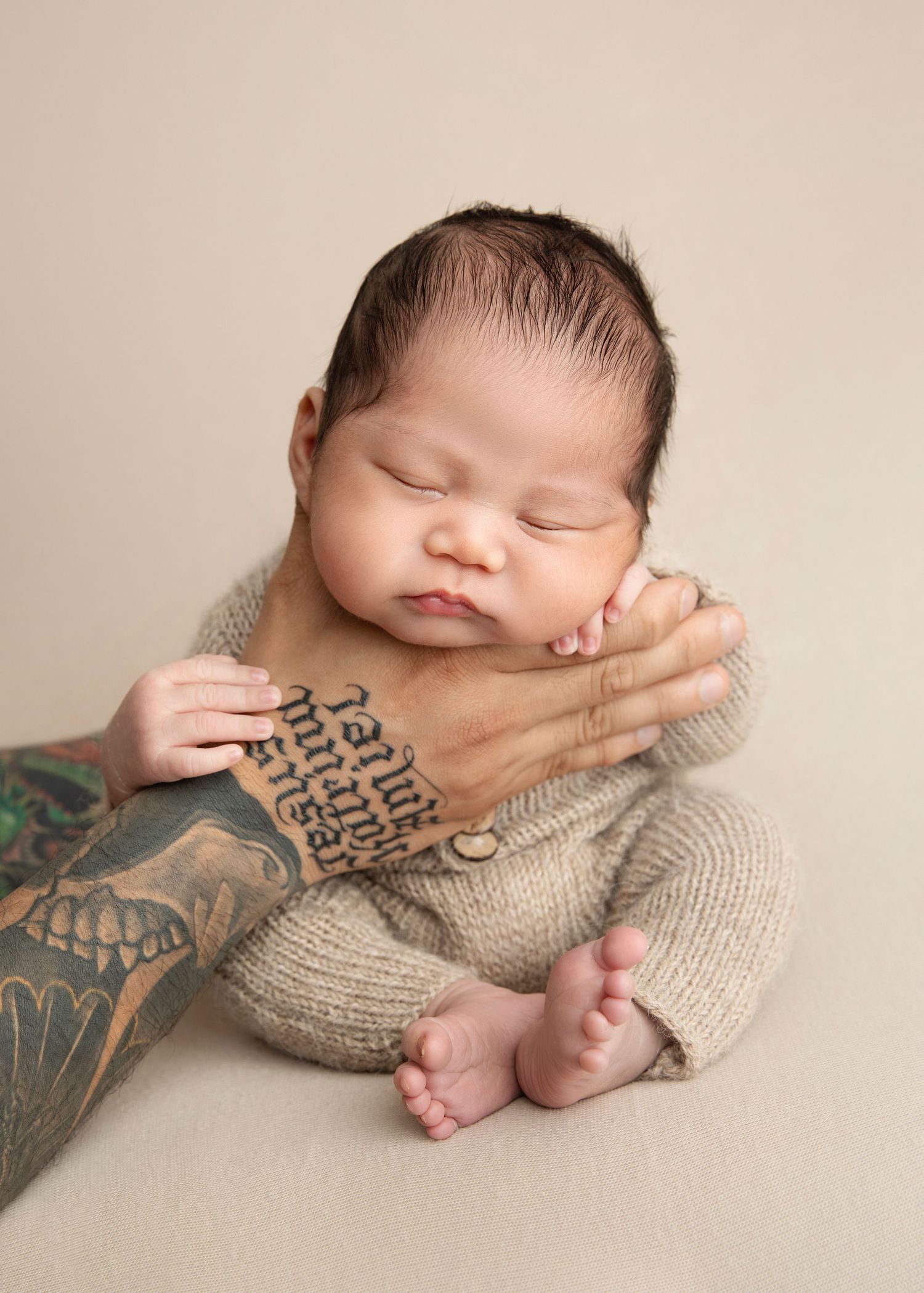 Sleeping newborn baby in beige knit outfit cradled by tattooed adult hand against neutral background.