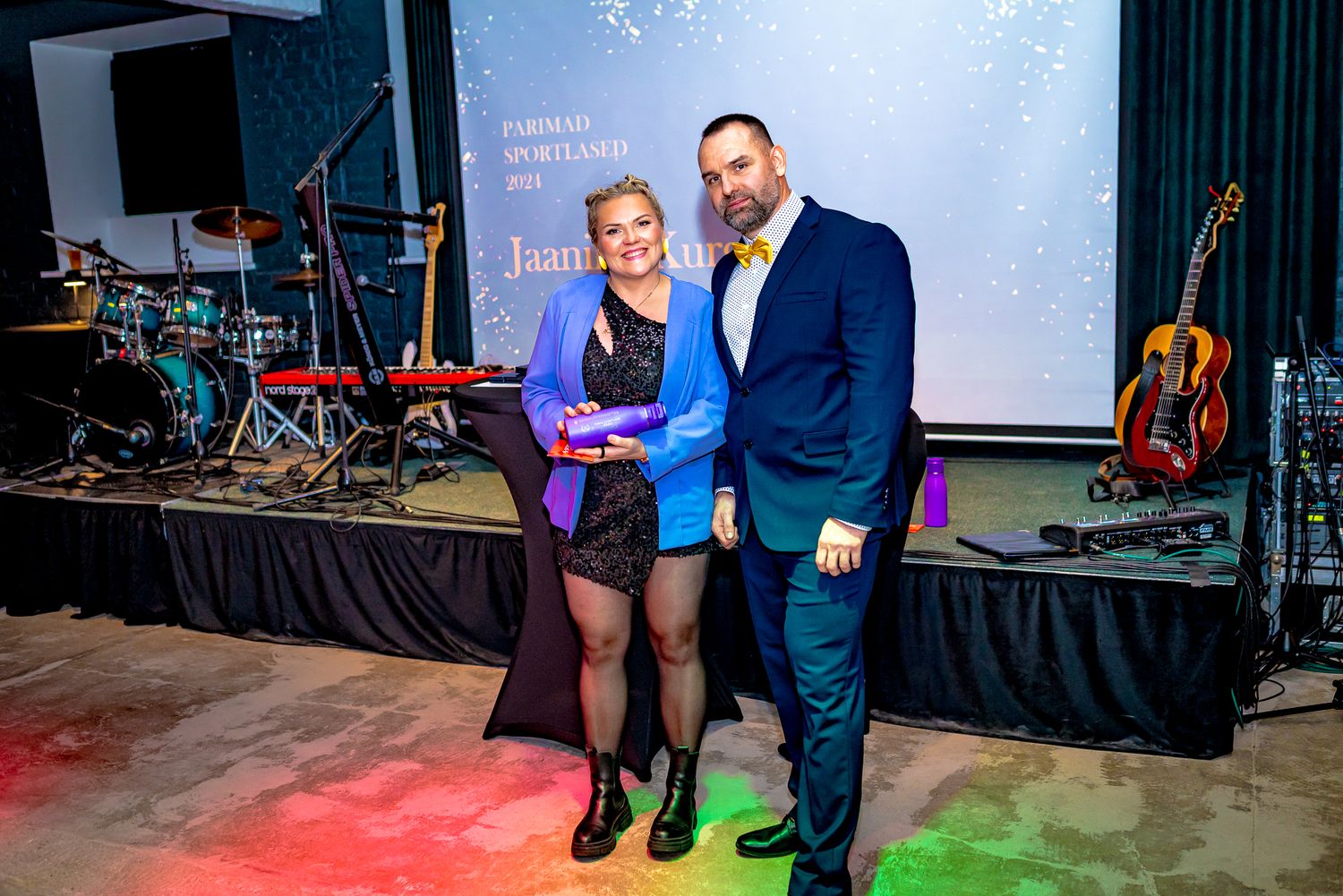 Two people posing with an award trophy on a colorful lit stage at a formal sports spotlight ceremony.