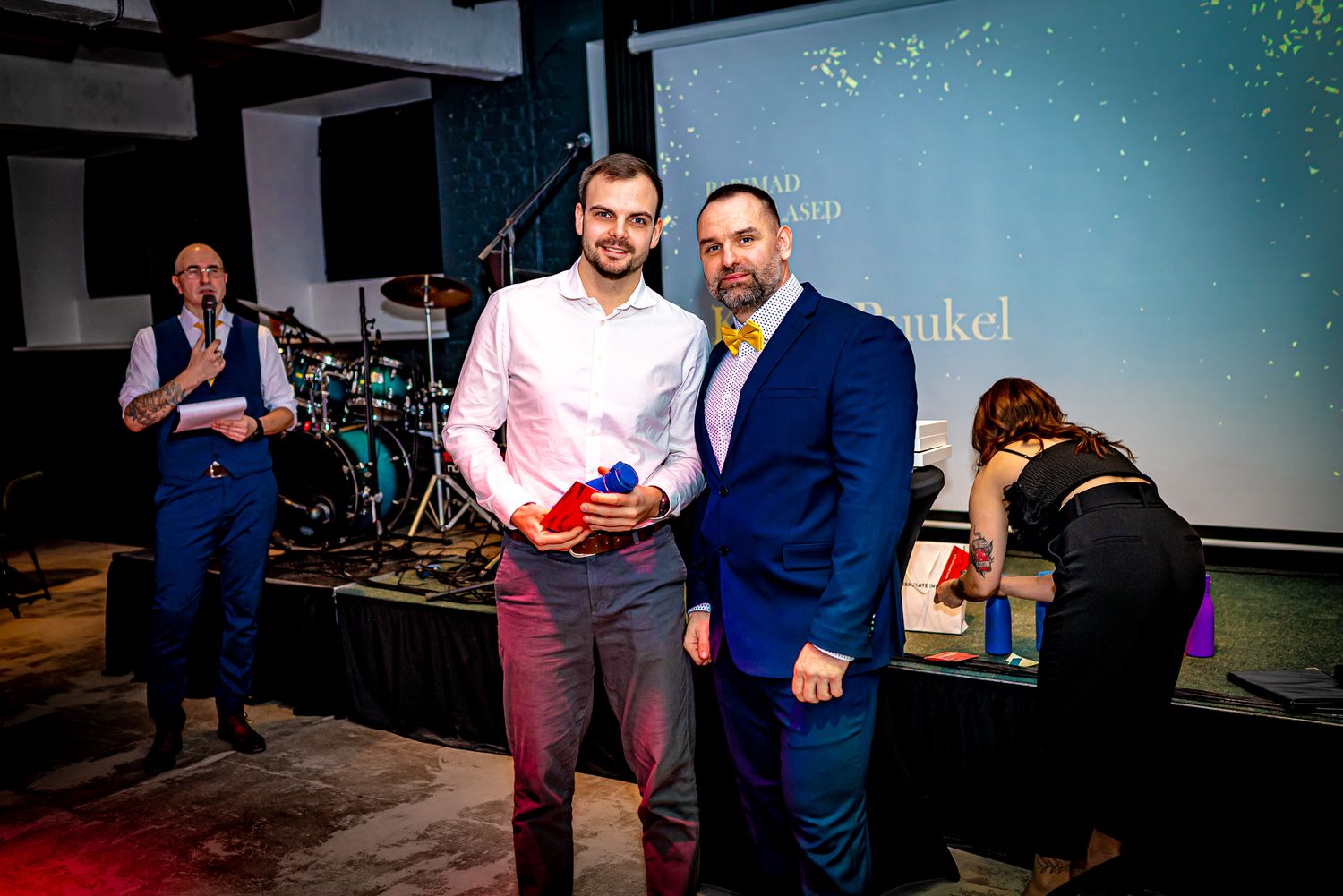 Two men pose together at an awards ceremony, one holding a trophy, with a live band performing on stage behind them.
