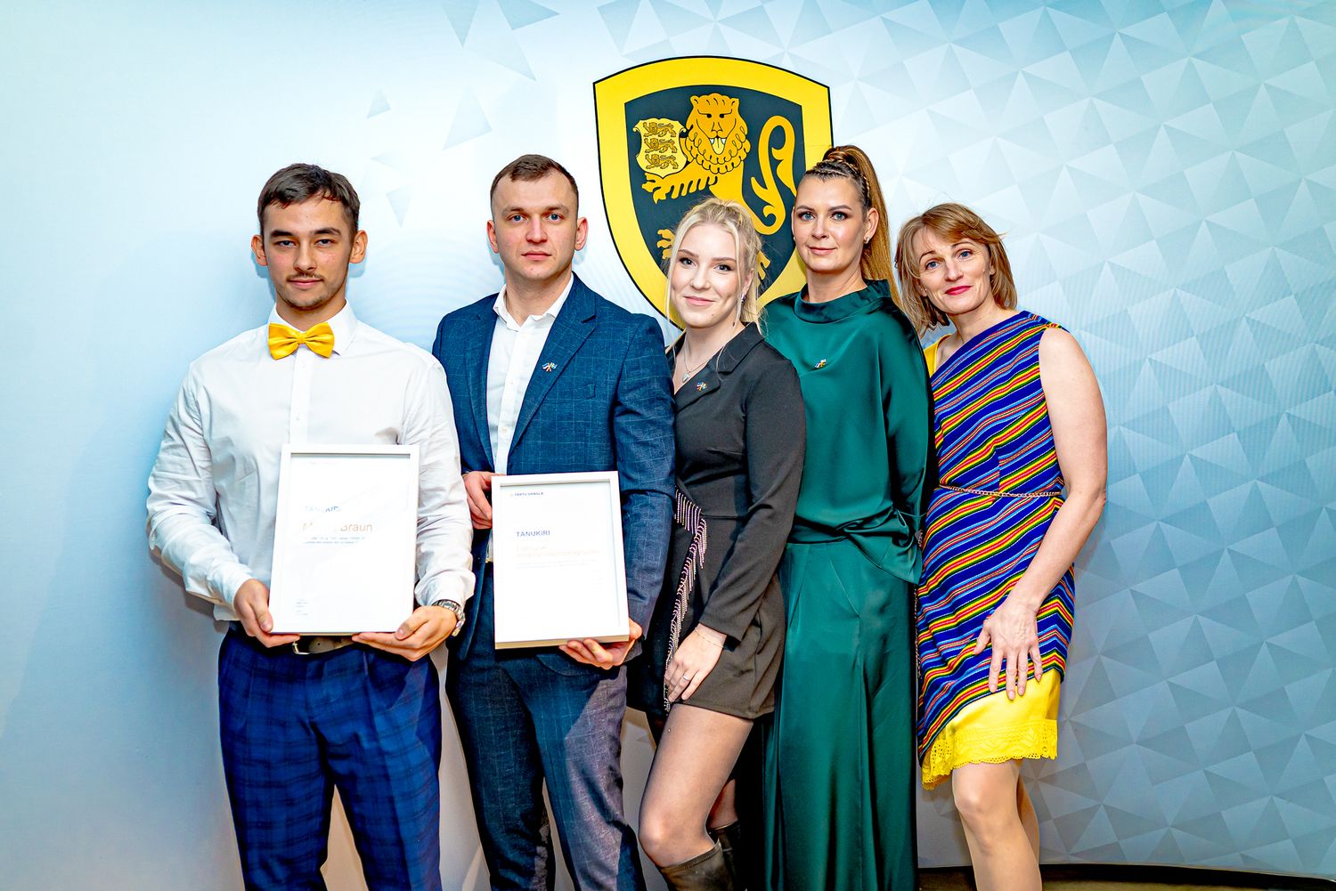 Five professionals posing with certificates at a formal recognition event before a golden crest backdrop.