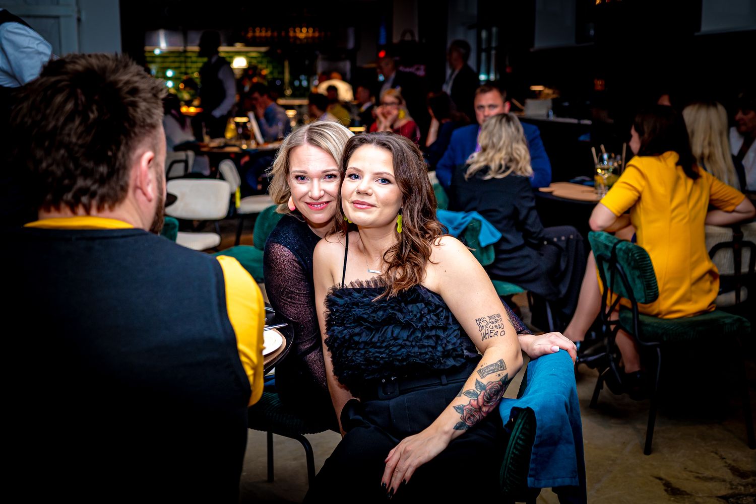 Two women smiling and posing together at a social event in a dimly lit venue with guests in the background.