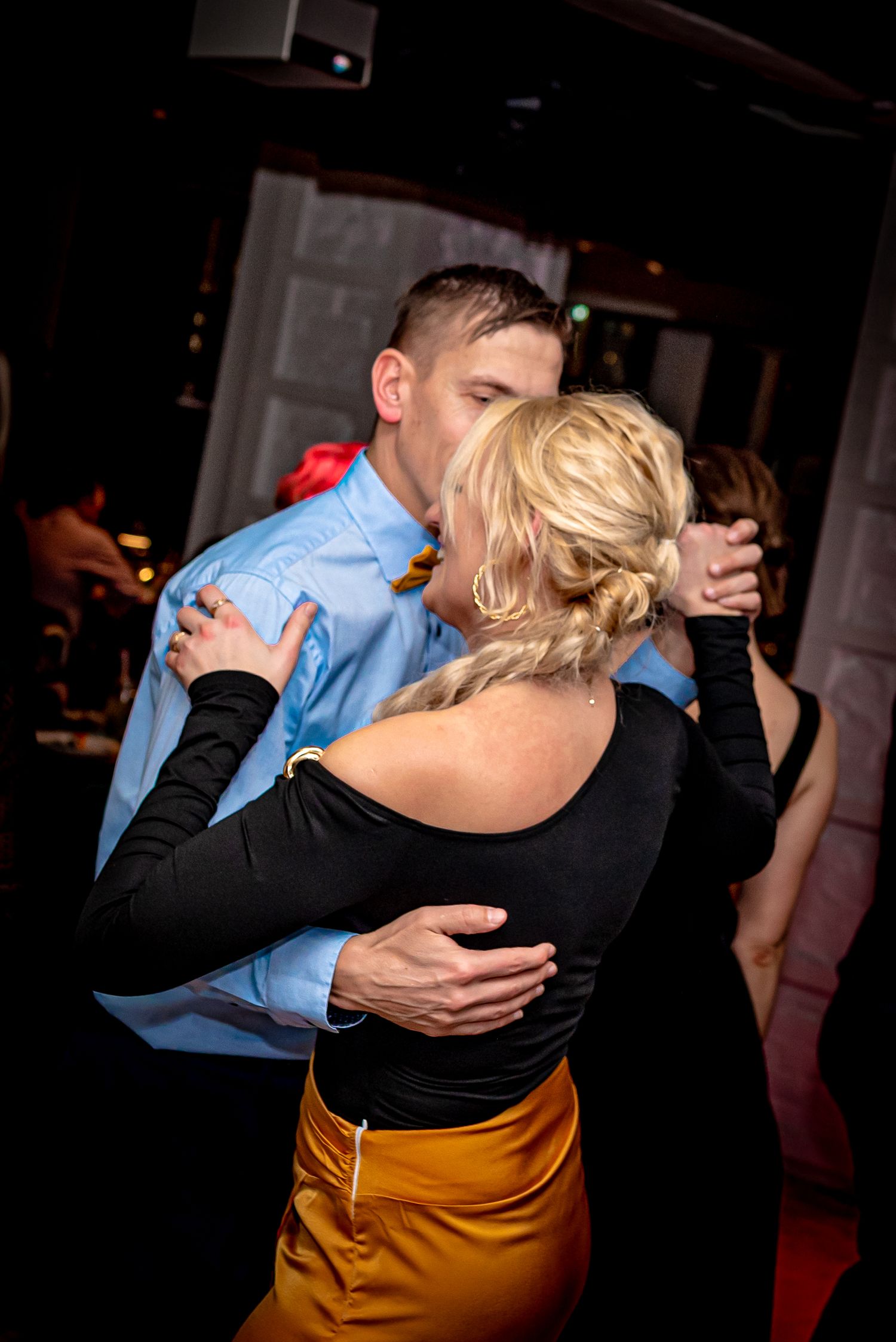 Couple slow dancing closely at a social event, man in blue shirt embracing woman in black top and gold skirt.