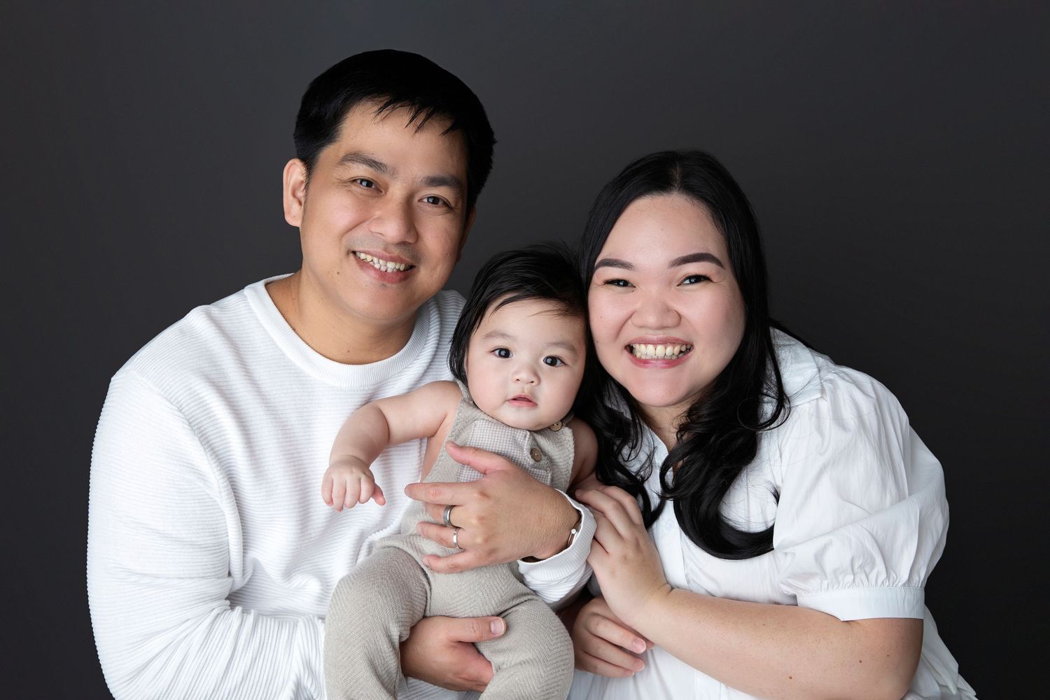 Happy Asian family portrait with smiling parents holding their baby, dressed in white, against a dark background.